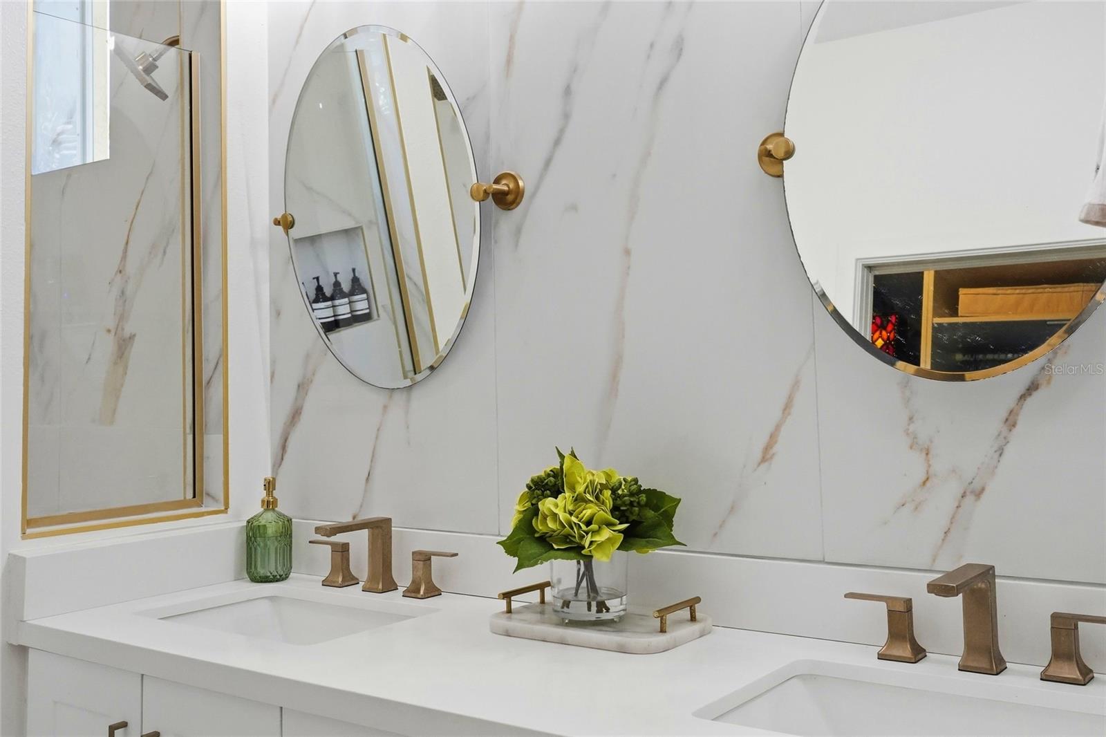 Bathroom vanity with double sinks, brass faucets, round mirrors, and veined marble-look walls.