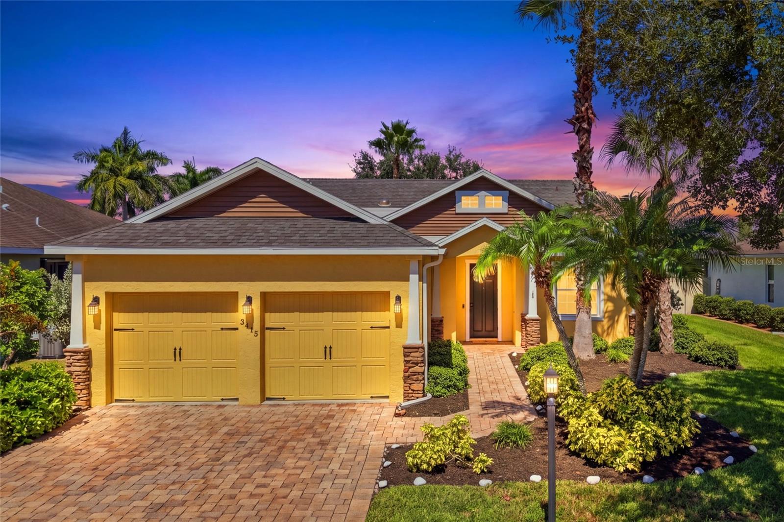 Yellow home with a two-car garage, brick driveway, palm trees, and landscaped front yard at dusk.