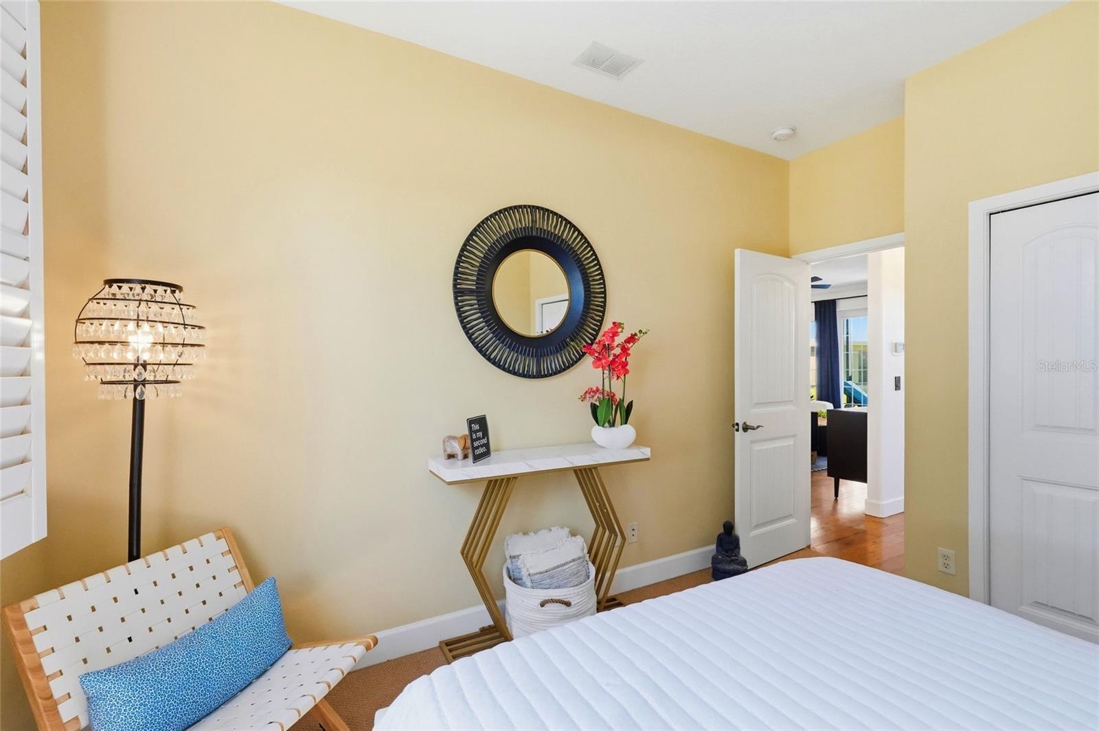 Bedroom with a white bed, yellow walls, woven chair, and round black mirror.