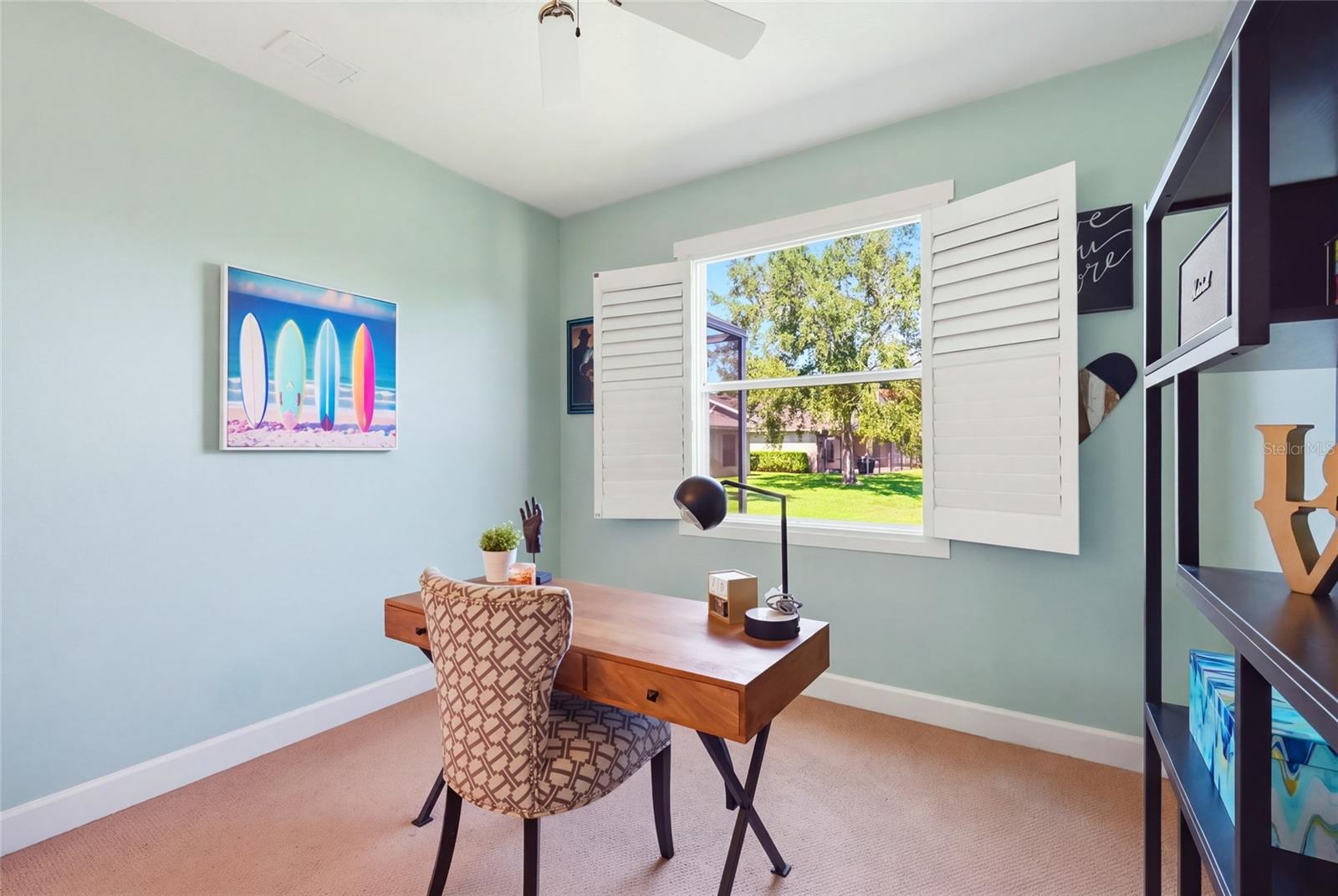 Home office with a wooden desk and shuttered window overlooking a sunny yard.