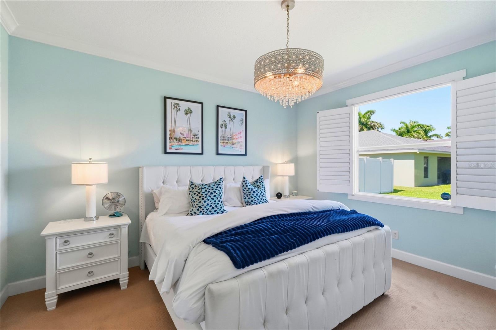 Staged bedroom with a white upholstered bed, blue accents, and a chandelier beside a shuttered window.