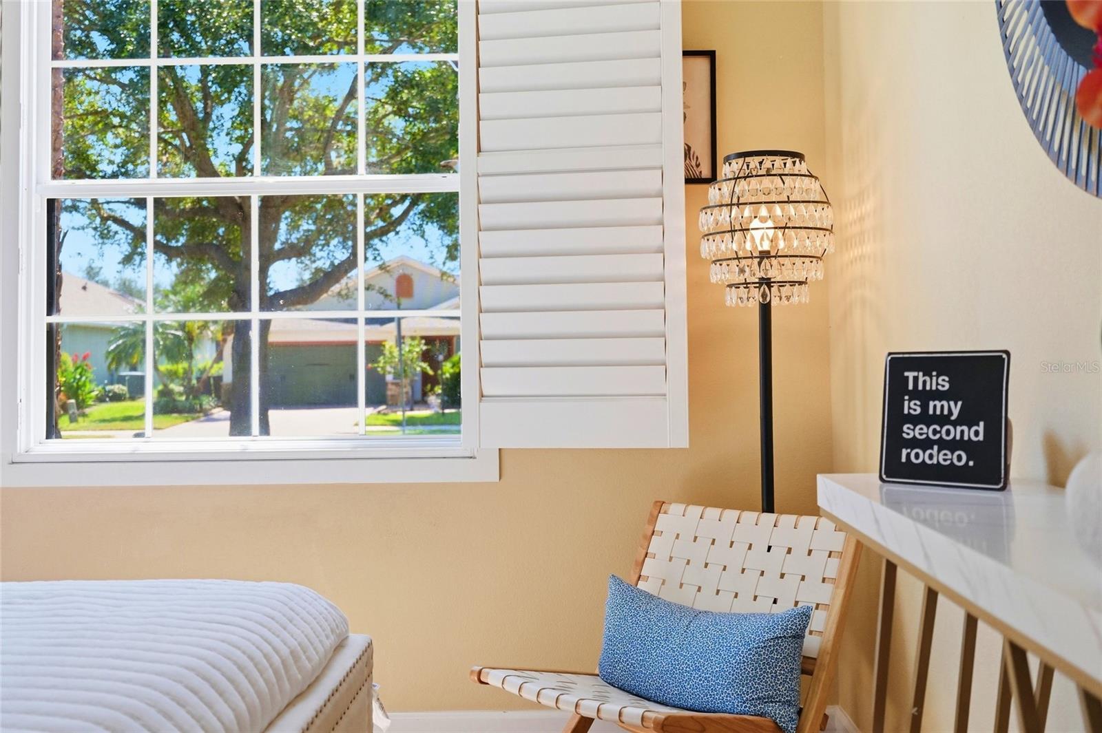 Bedroom with a shuttered window, woven chair, blue cushion, and a sign reading “This is my second rodeo.”