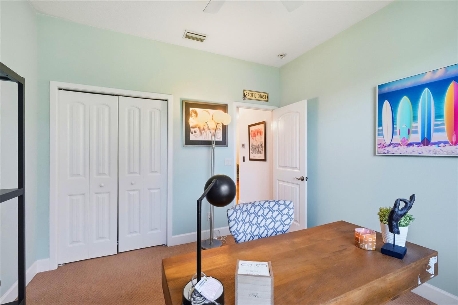 Furnished home office with a wooden desk, white closet, and colorful surfboard artwork.