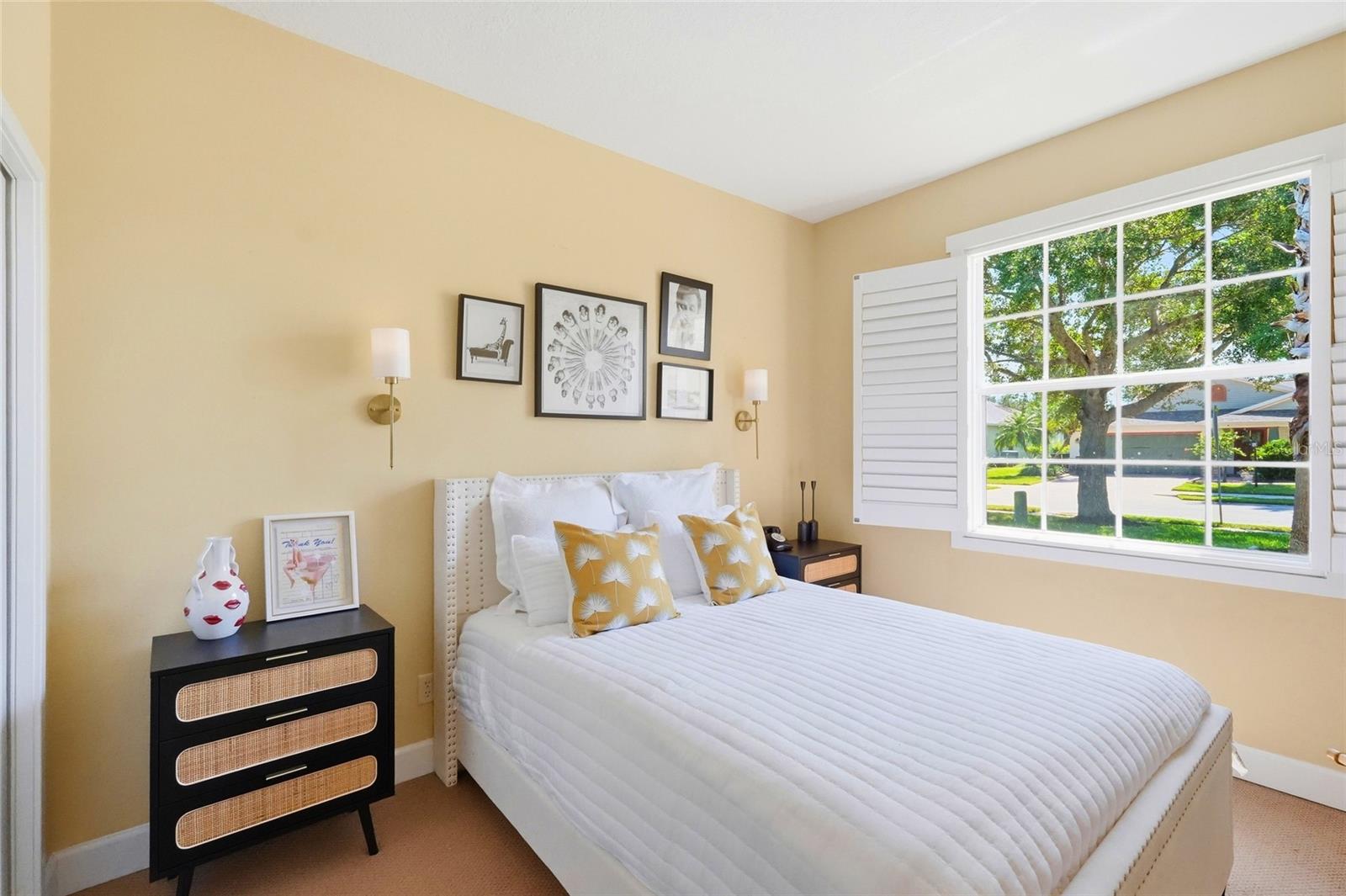 Bedroom with a white bed, mustard-yellow pillows, and a large window overlooking a leafy street.
