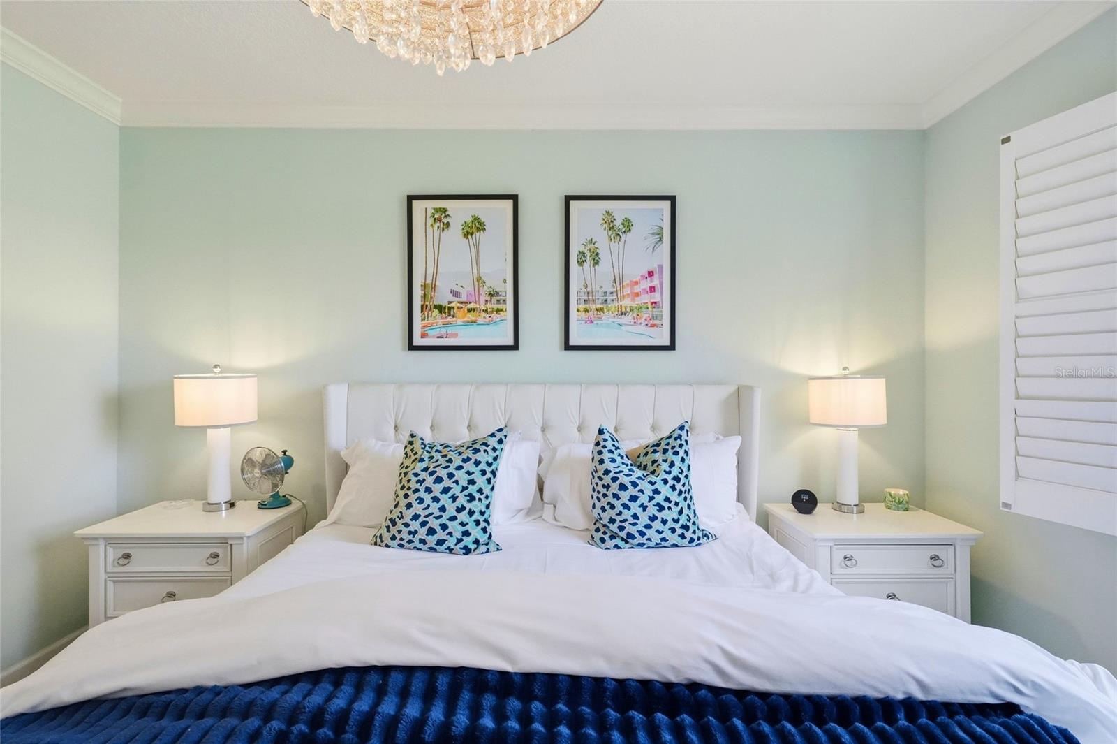 Furnished bedroom with a white bed, blue patterned pillows, mint-green walls, and framed palm-tree prints.