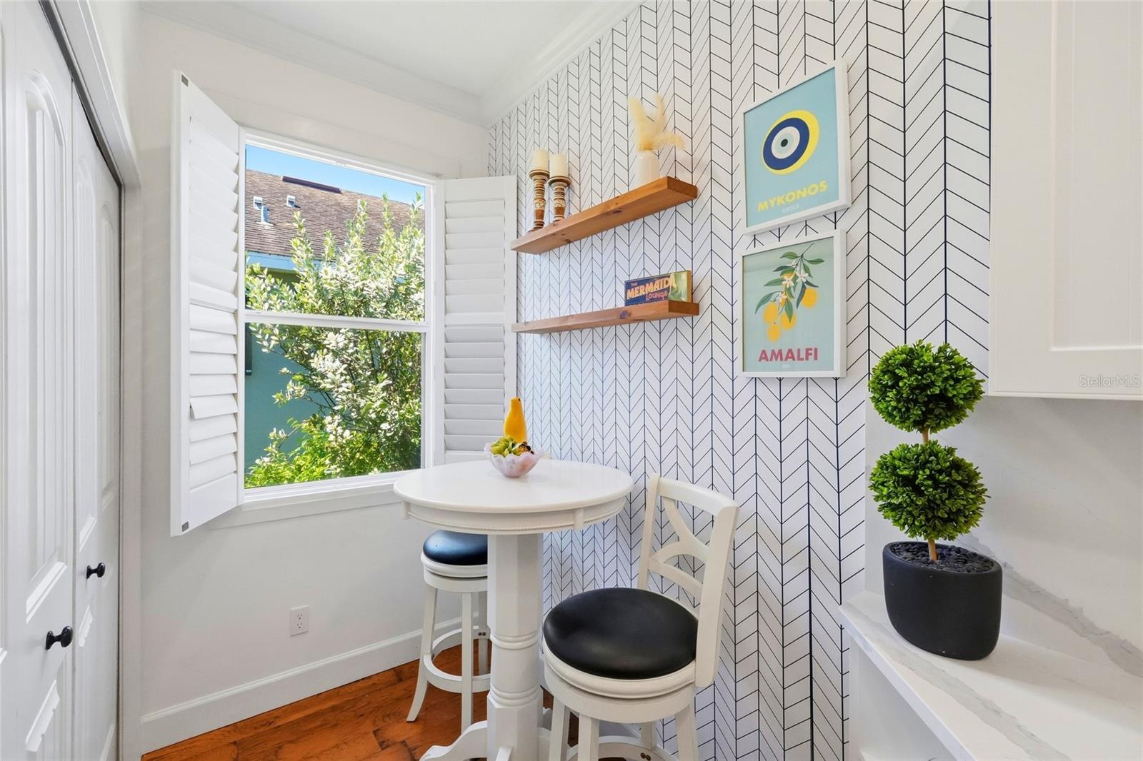 Breakfast nook with a round white table, shuttered window, geometric wallpaper, and wood shelves.