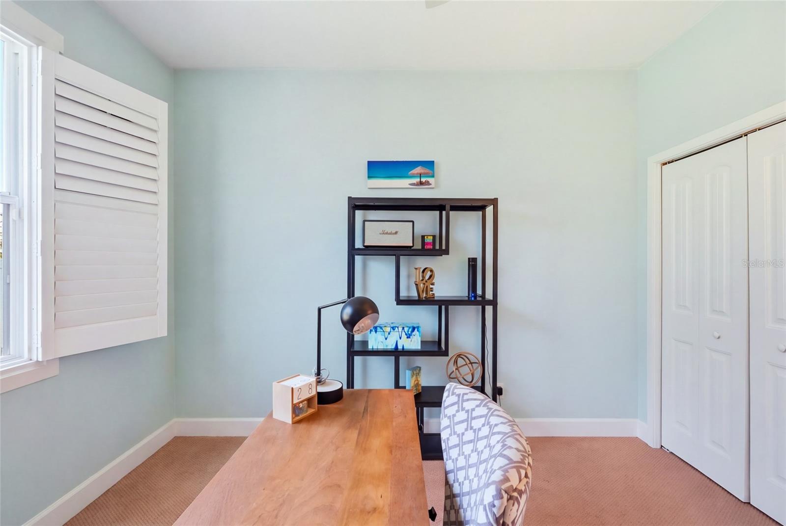 Furnished home office with a wooden desk, pale aqua walls, and white plantation shutters.
