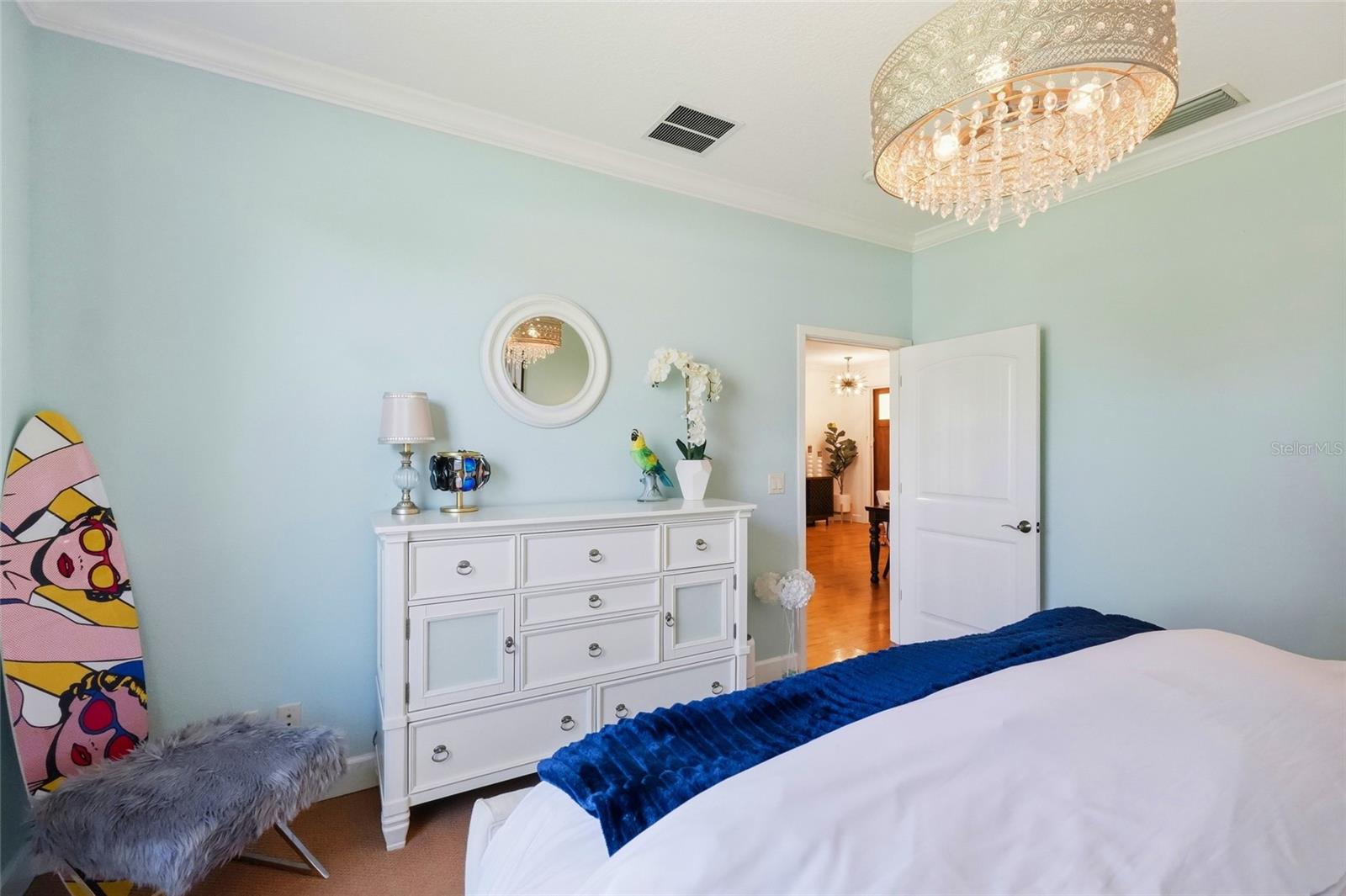 Bedroom with pale blue walls, a crystal chandelier, white dresser, and colorful surfboard.