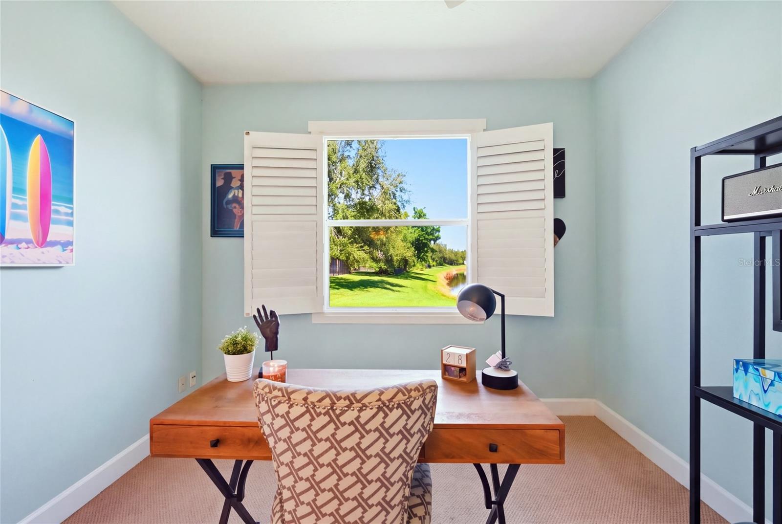 Home office with a wooden desk and shuttered window overlooking a green lawn and water.