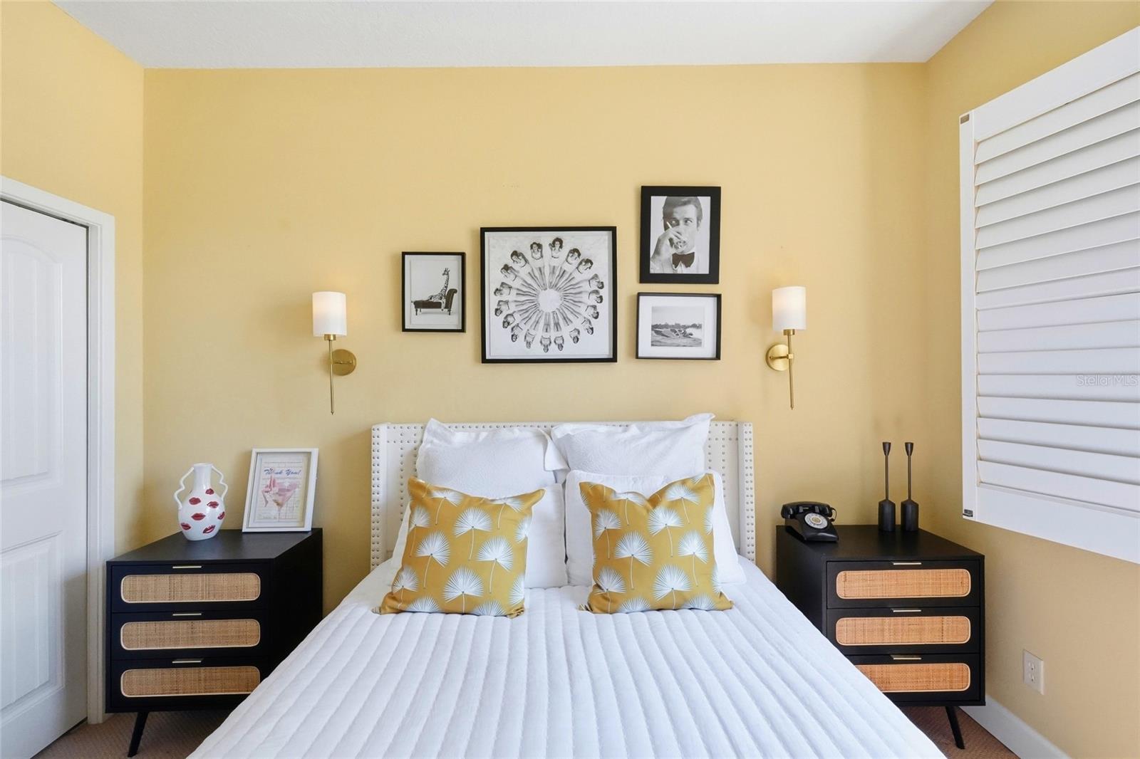 Furnished bedroom with a white bed, mustard-yellow pillows, framed artwork, and matching black nightstands.
