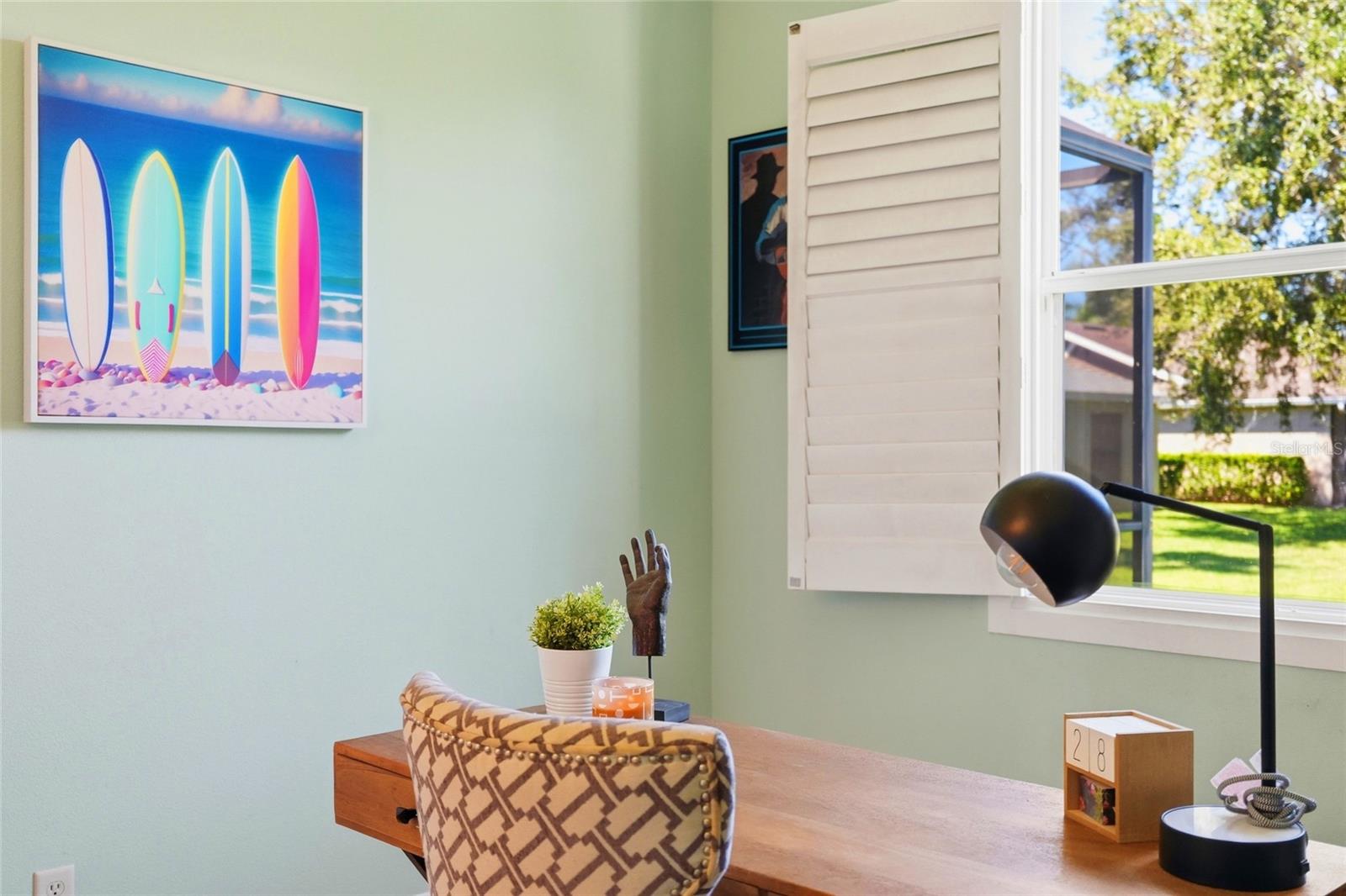 Home office with a wooden desk, patterned chair, white shutters, and colorful surfboard artwork.
