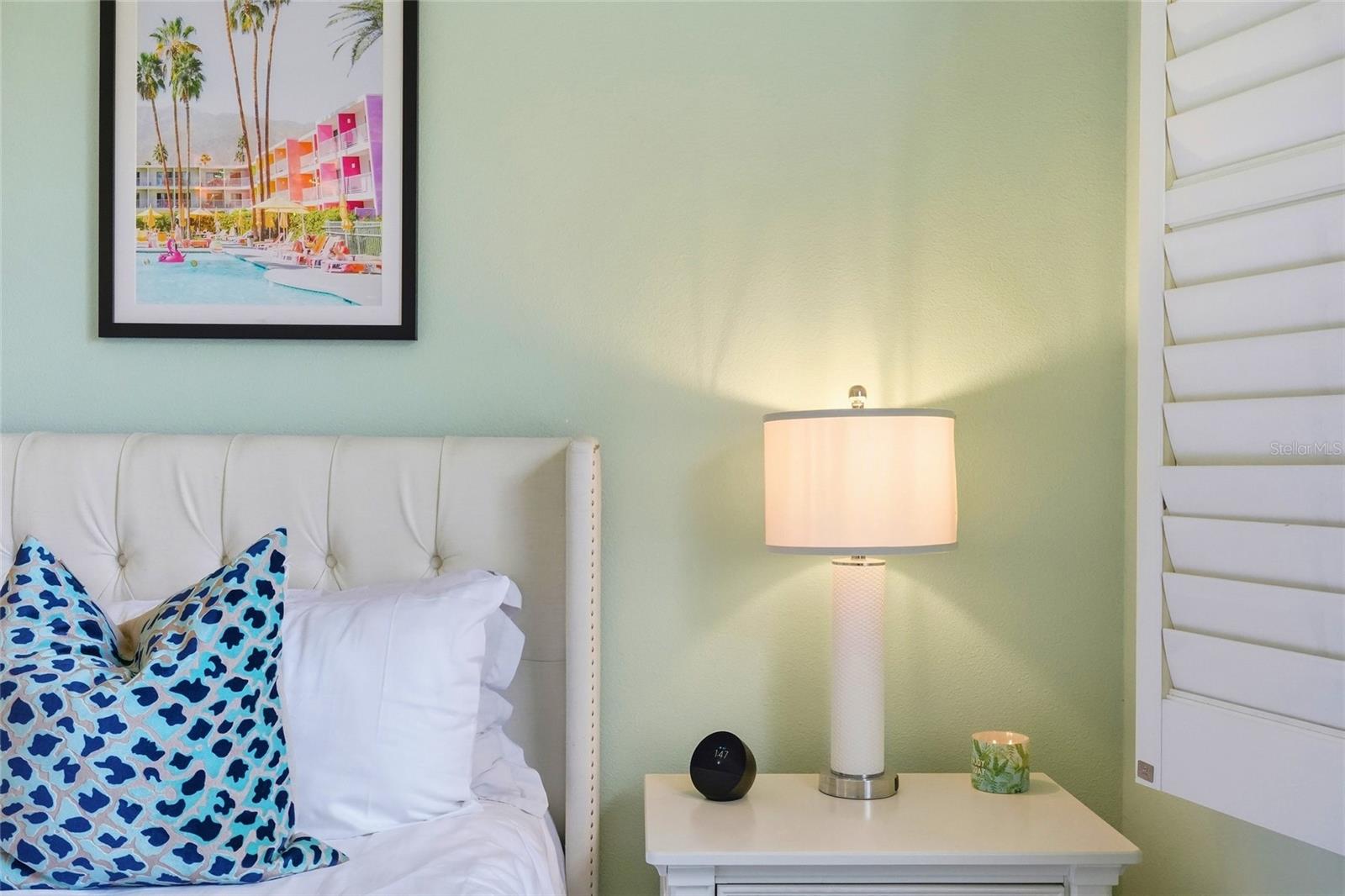 Bedroom bedside with a white upholstered bed, blue patterned pillows, lamp, and framed poolside artwork.