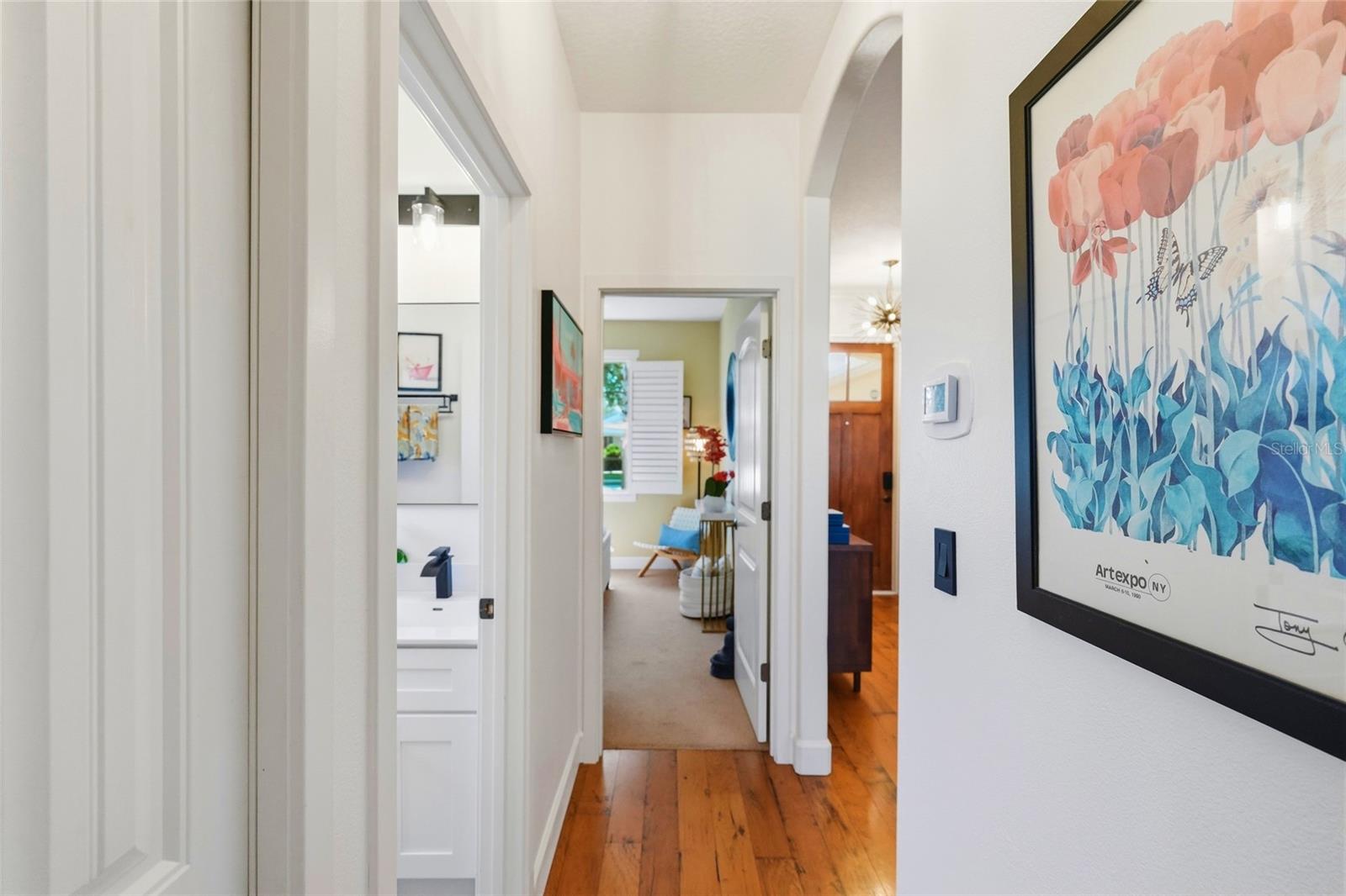 Bright hallway with warm wood floors, framed floral artwork, and open doorways to adjoining rooms.