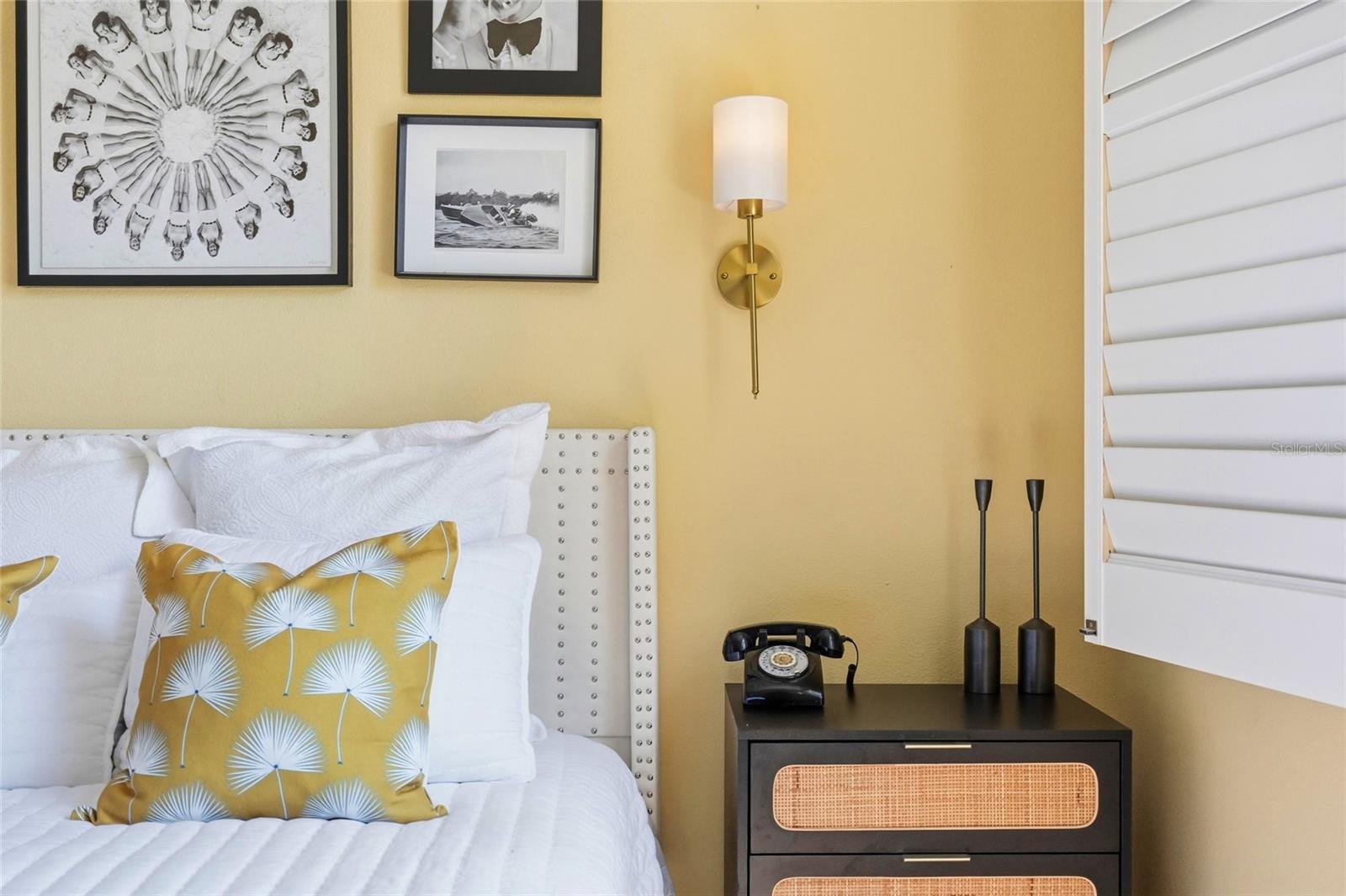 Bedroom with mustard-yellow accent pillows, framed black-and-white art, and a black bedside chest.