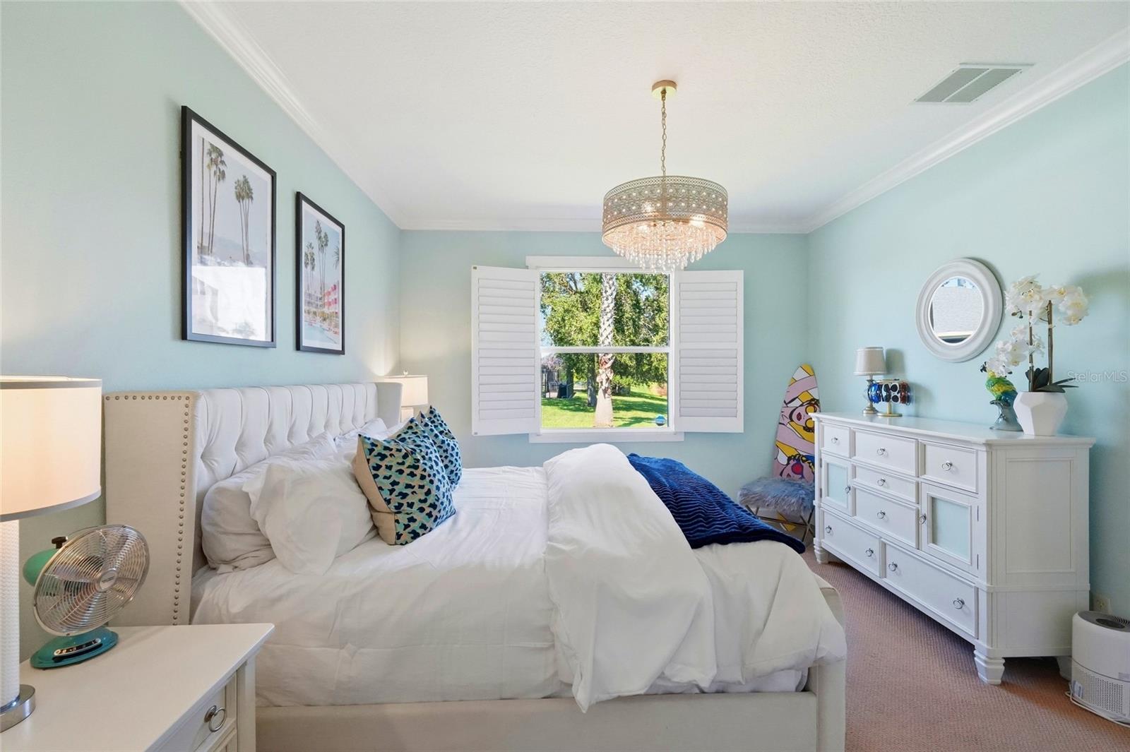Bedroom with a white upholstered bed, crystal chandelier, and shuttered window overlooking greenery.