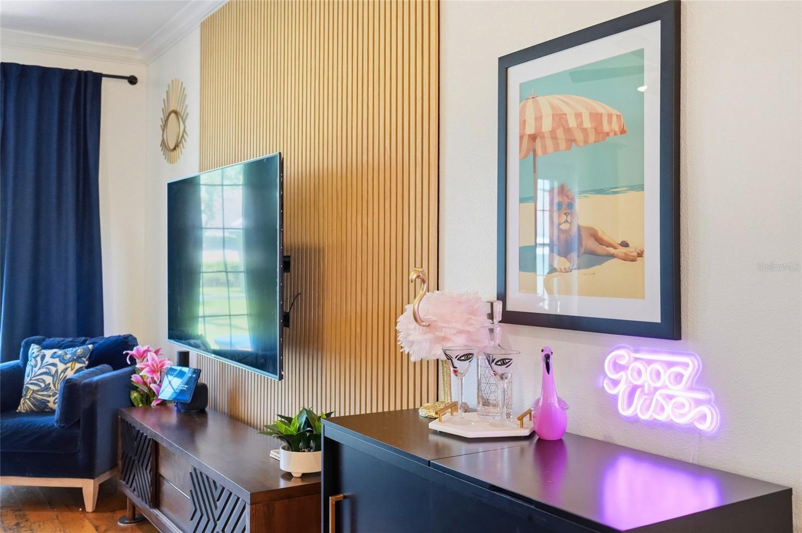 Living room with a navy sofa, wood-slat TV wall, beach artwork, and glowing “Good Vibes” sign.