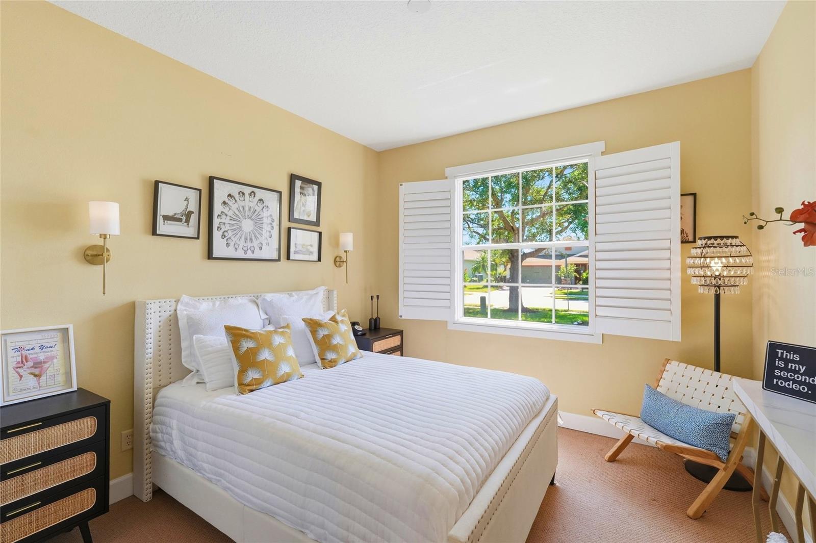 Bedroom with a white bed, mustard-yellow pillows, and open plantation shutters overlooking a leafy street.