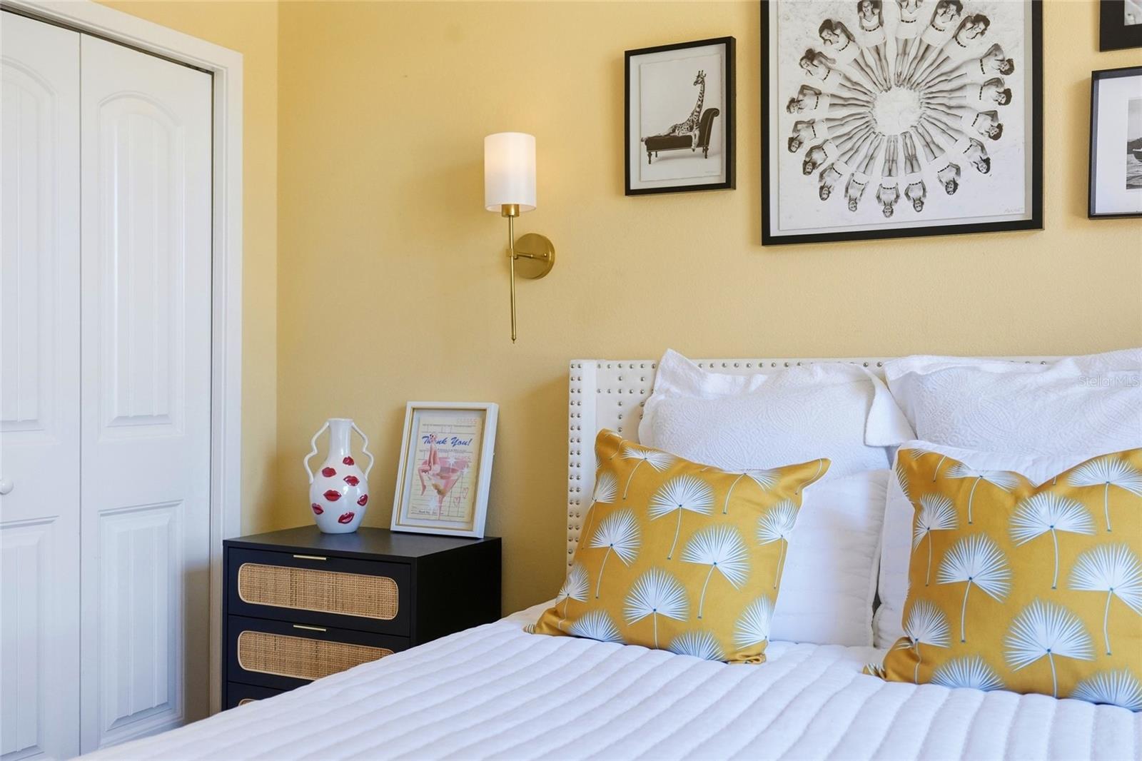 Bedroom with a white bed, mustard patterned pillows, yellow walls, and framed artwork.