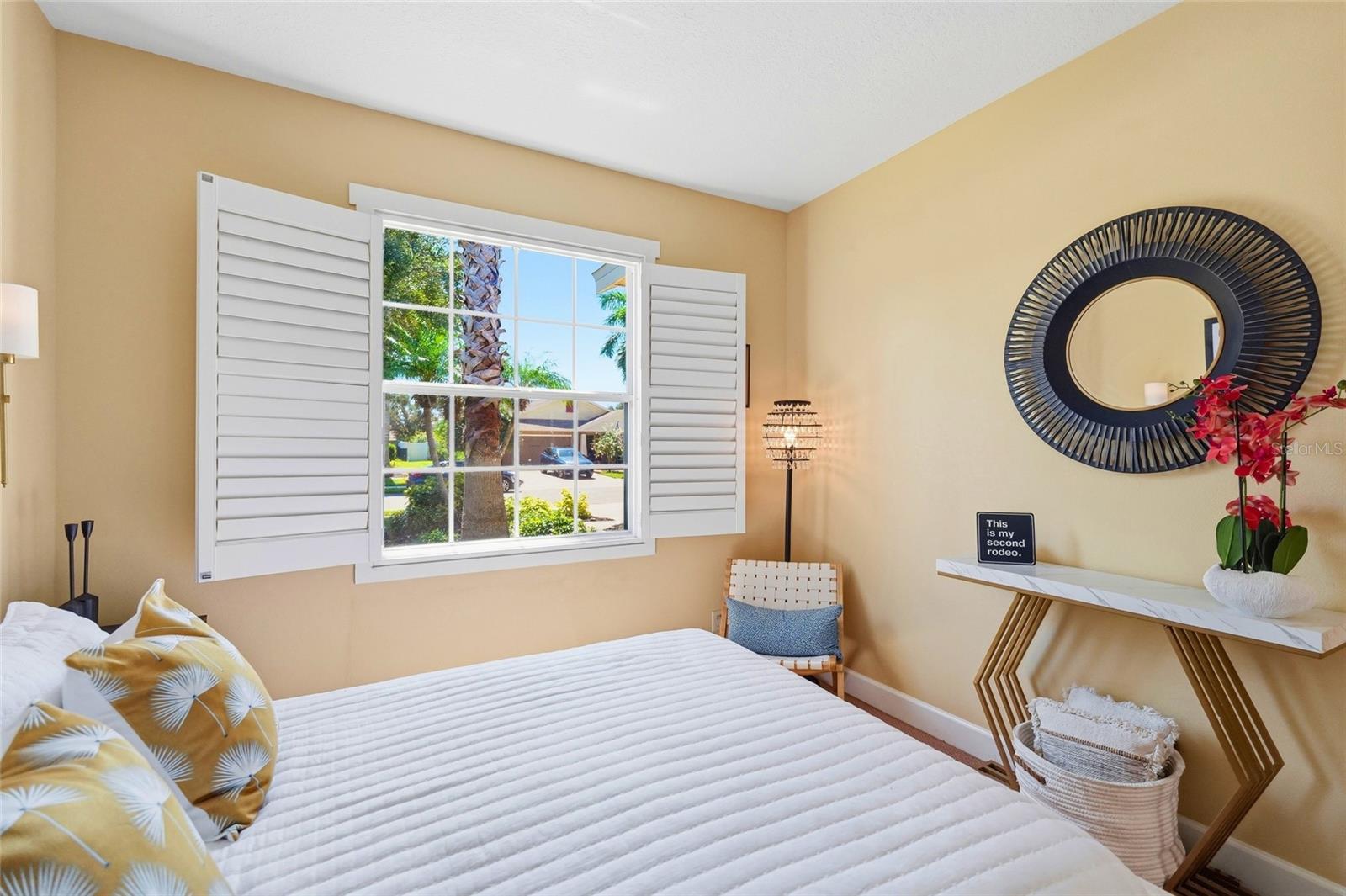 Bedroom with a white quilted bed, yellow patterned pillows, and shuttered window overlooking palm trees.