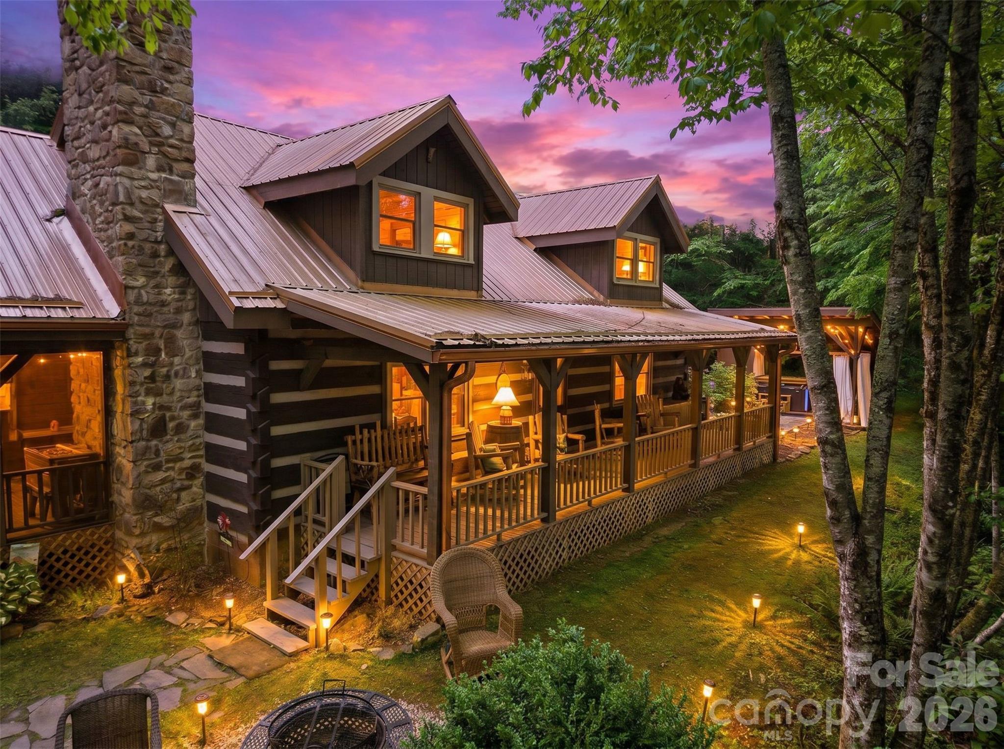 Rustic home with a stone chimney and illuminated covered porch beneath a pink-purple dusk sky.