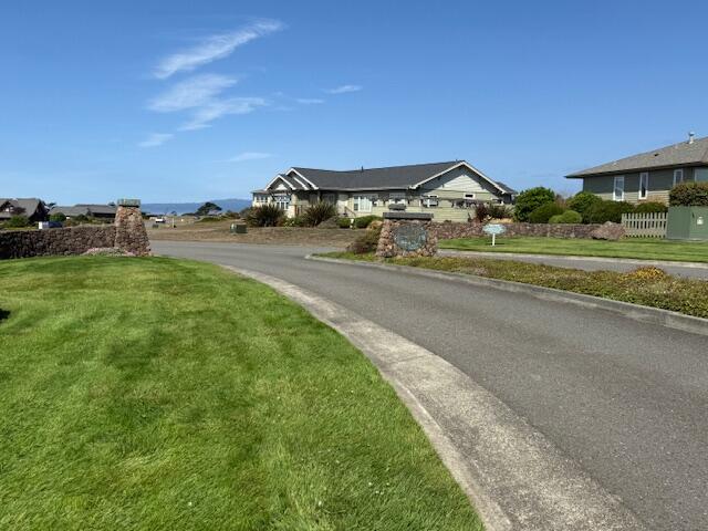 Single-story home beyond a green lawn and stone entrance pillars beside a curved residential road.