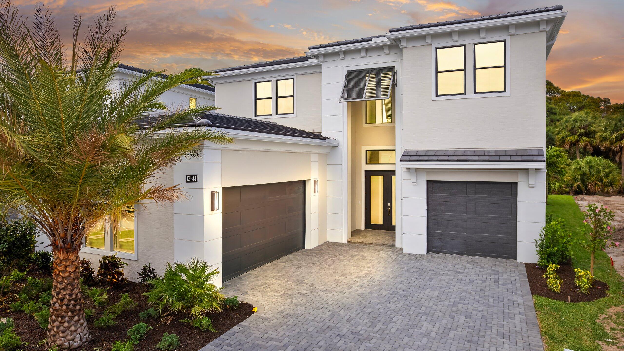 Modern two-story white home with dark double garage doors, a paver driveway, and palm trees at dusk.
