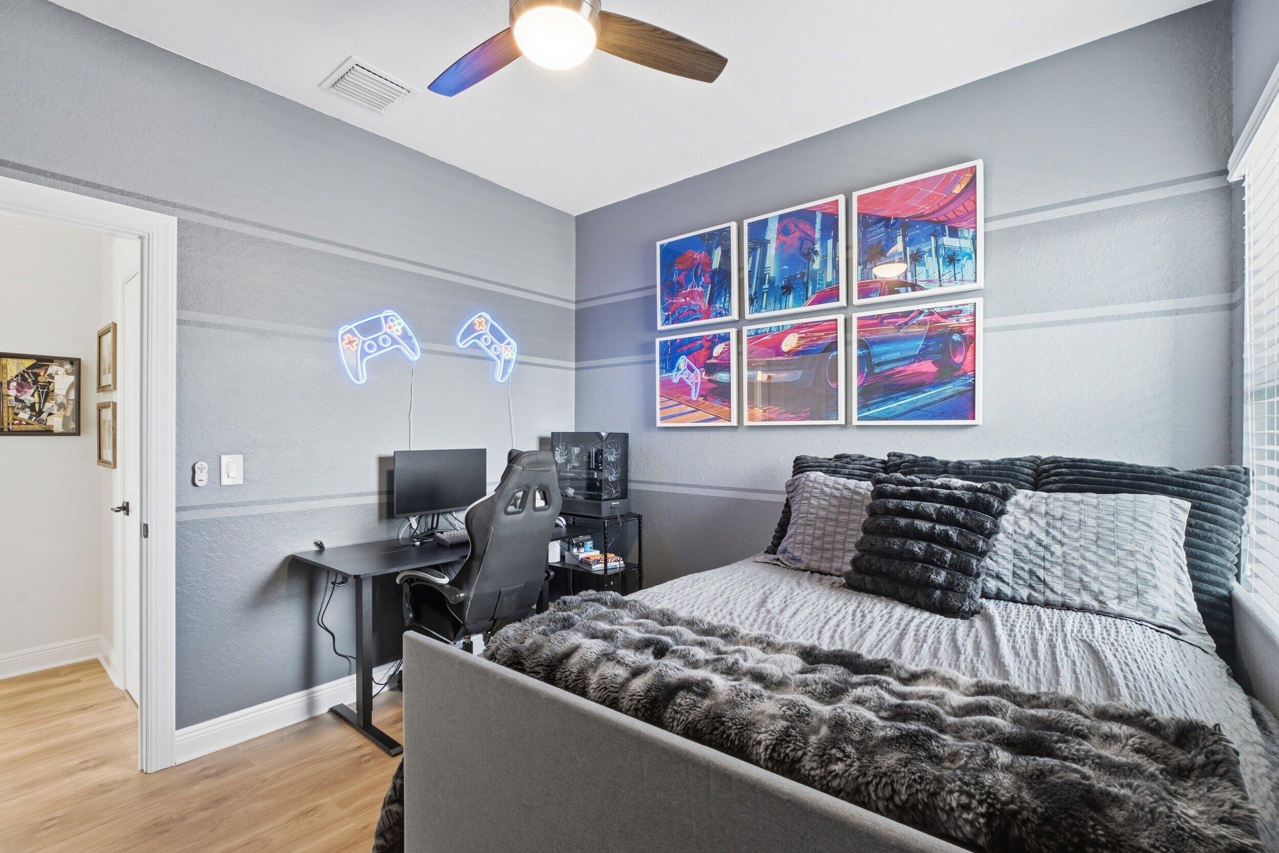 Bedroom with layered gray bedding, gaming desk, glowing controller lights, and colorful framed art.
