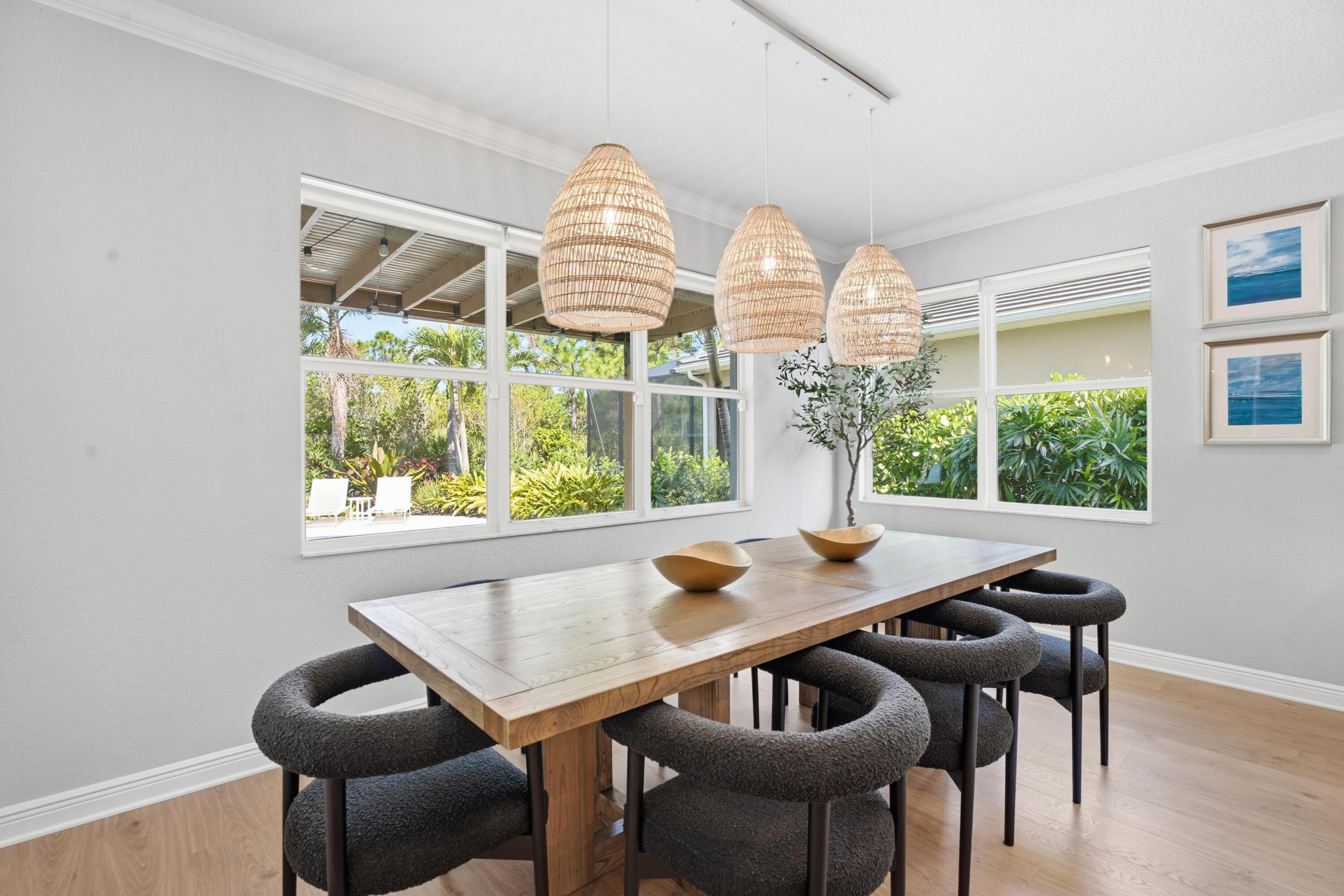 Dining room with a wooden table, dark chairs, woven pendant lights, and windows overlooking tropical greenery.