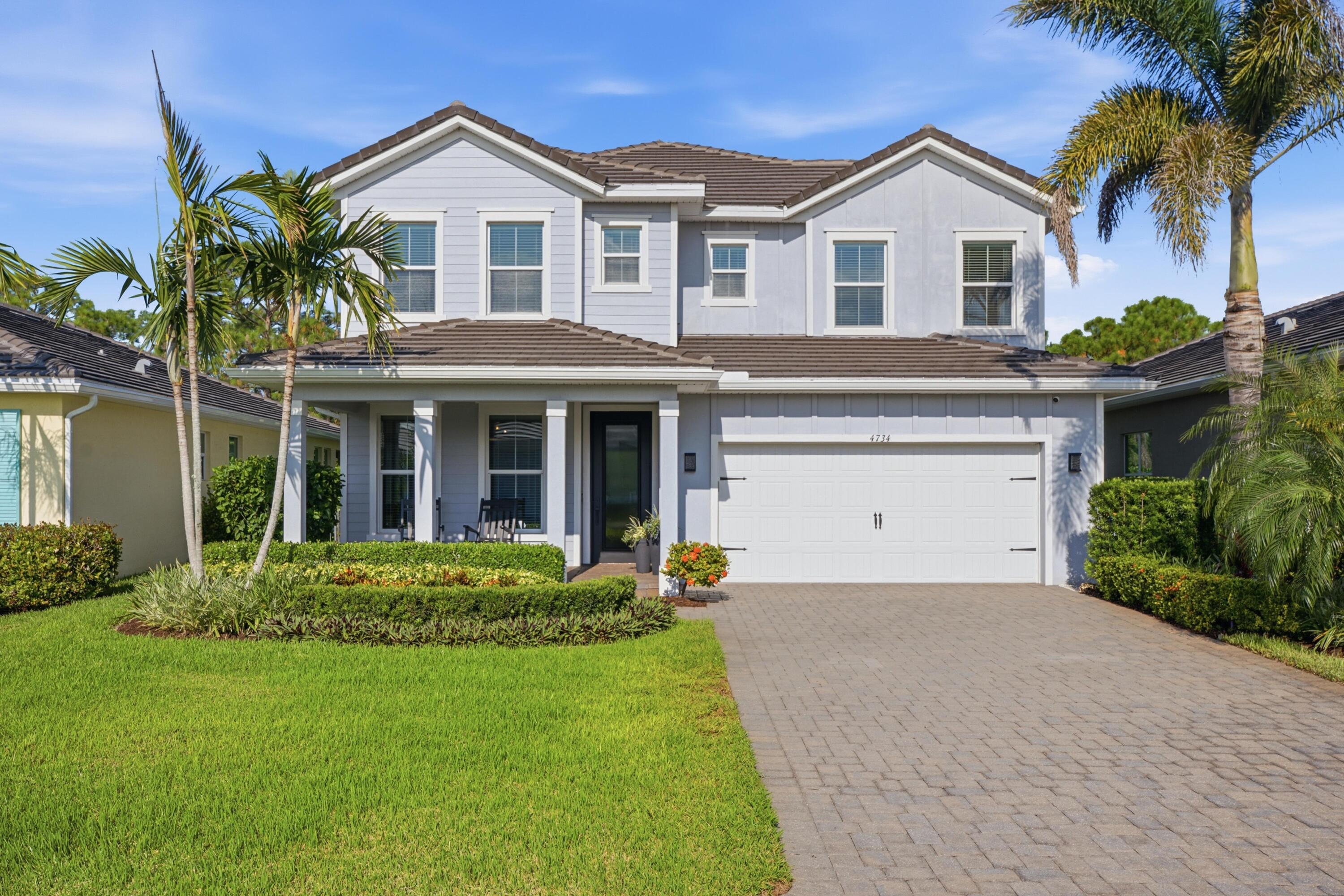 Two-story gray home with a tiled roof, two-car garage, paved driveway, and palm-lined landscaping.