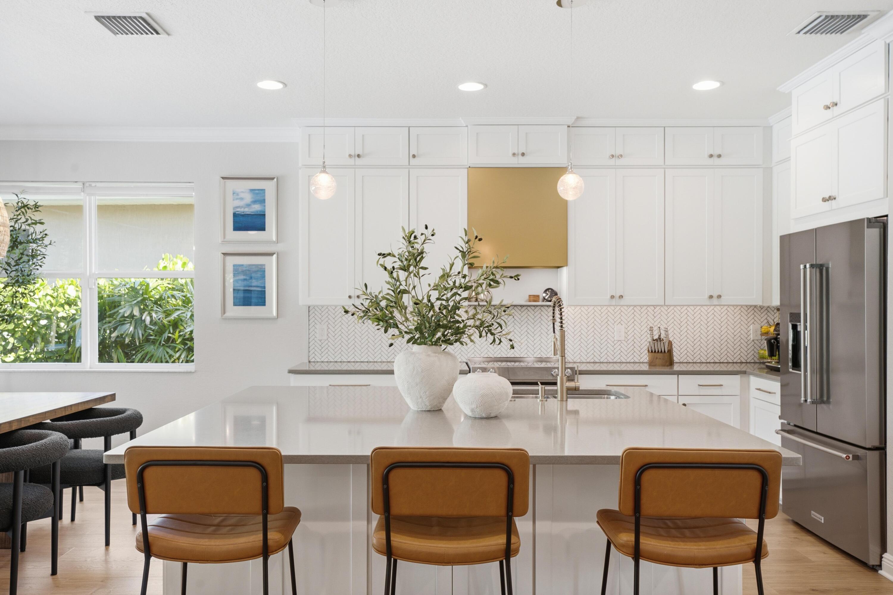 Bright white kitchen with a large island, three brown stools, herringbone backsplash, and garden-facing window.