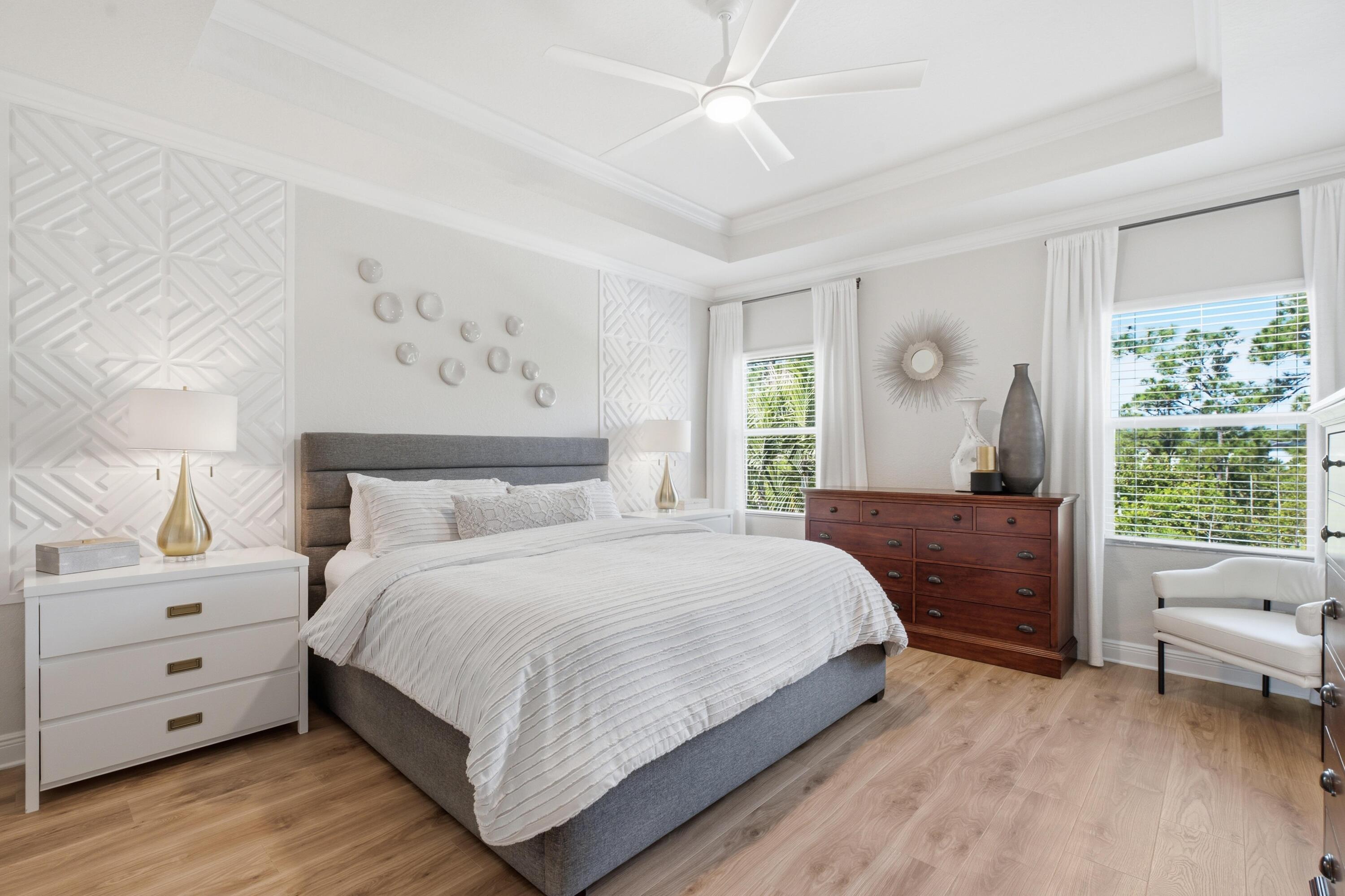 Bright bedroom with a gray bed, geometric wall panels, wood floors, and large curtained windows.
