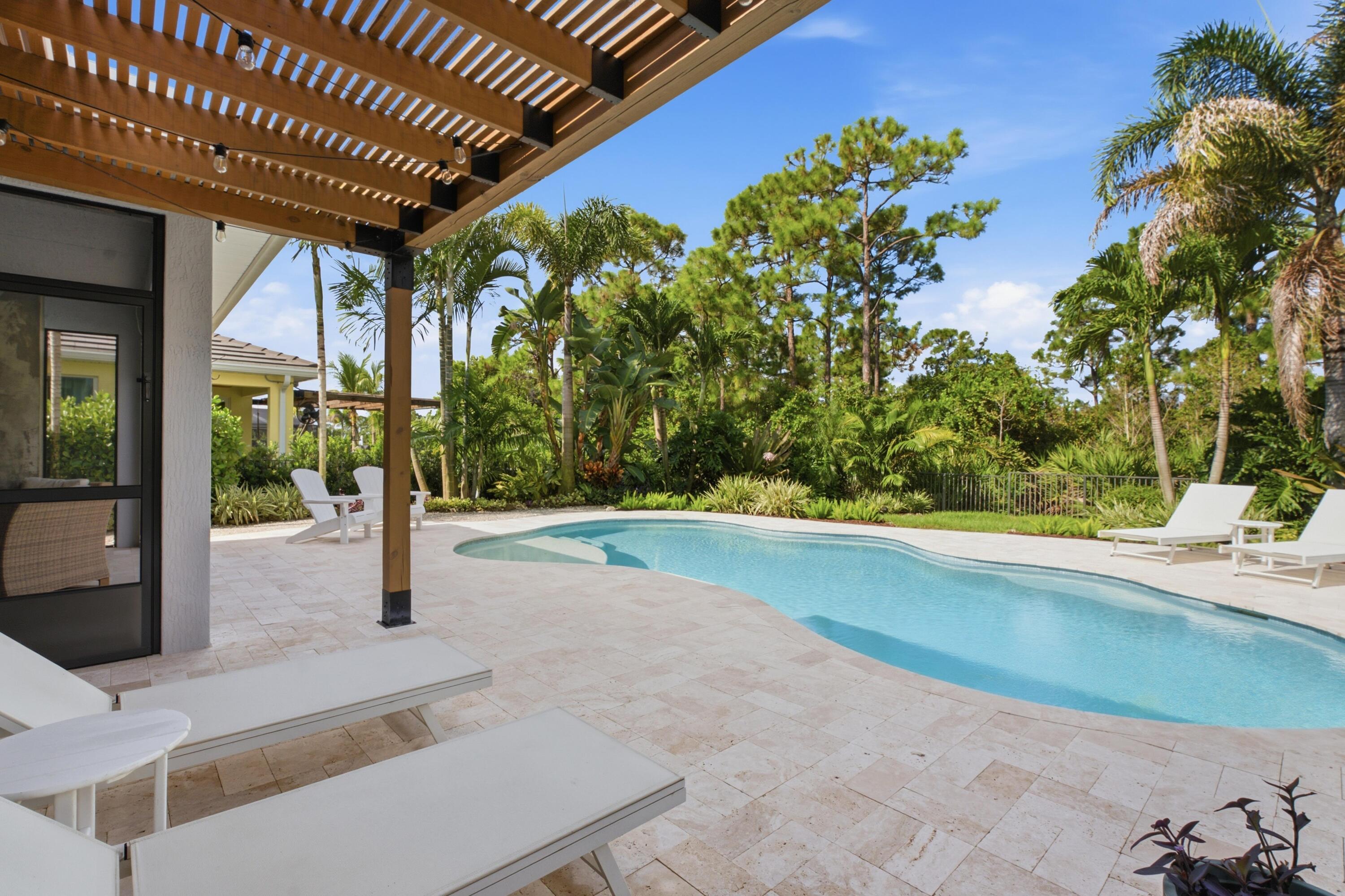 Curved backyard pool beneath a wooden pergola, surrounded by stone paving, lounge chairs, and tropical greenery.