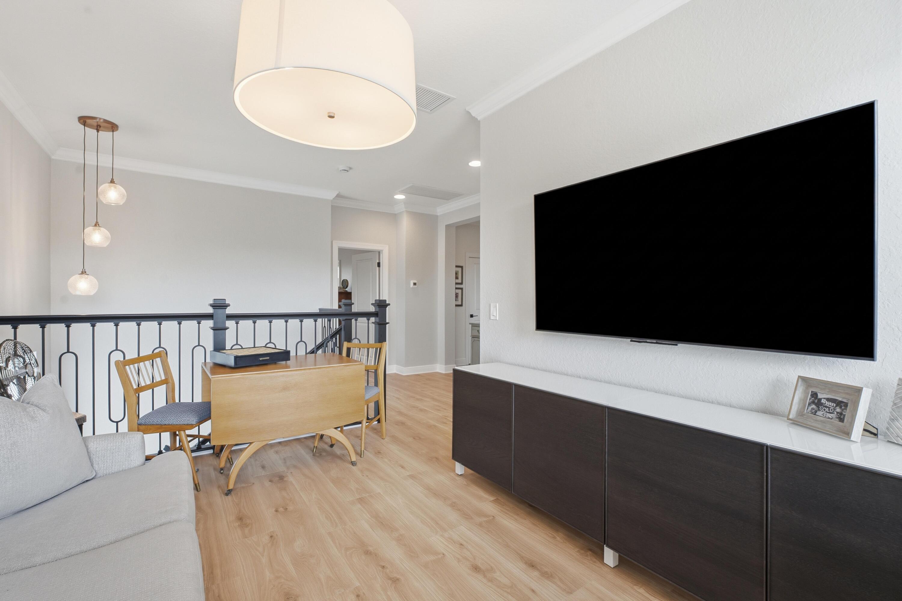 Upstairs living area with a gray sofa, dining table, black railing, and large wall-mounted television.