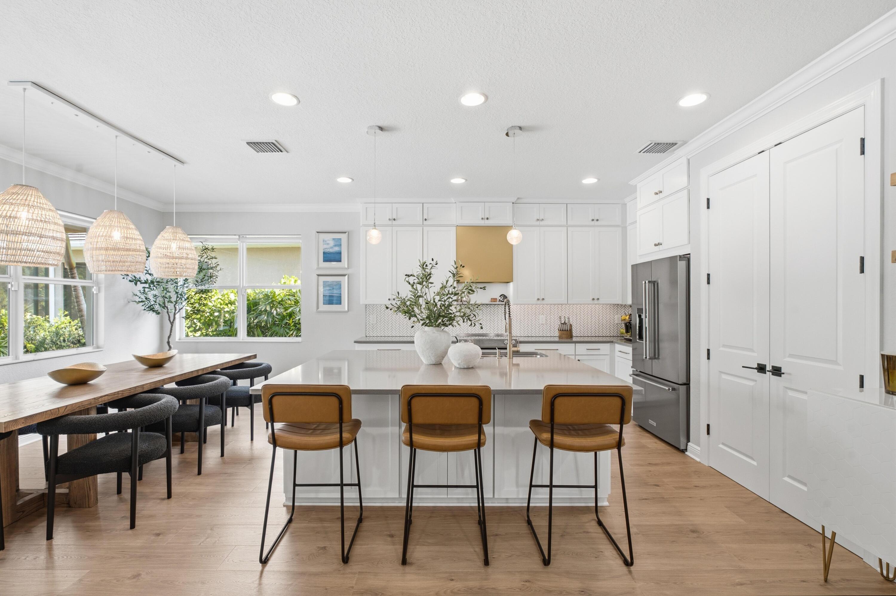 Bright white kitchen with a large island, three tan stools, woven pendant lights, and a window-side dining table.