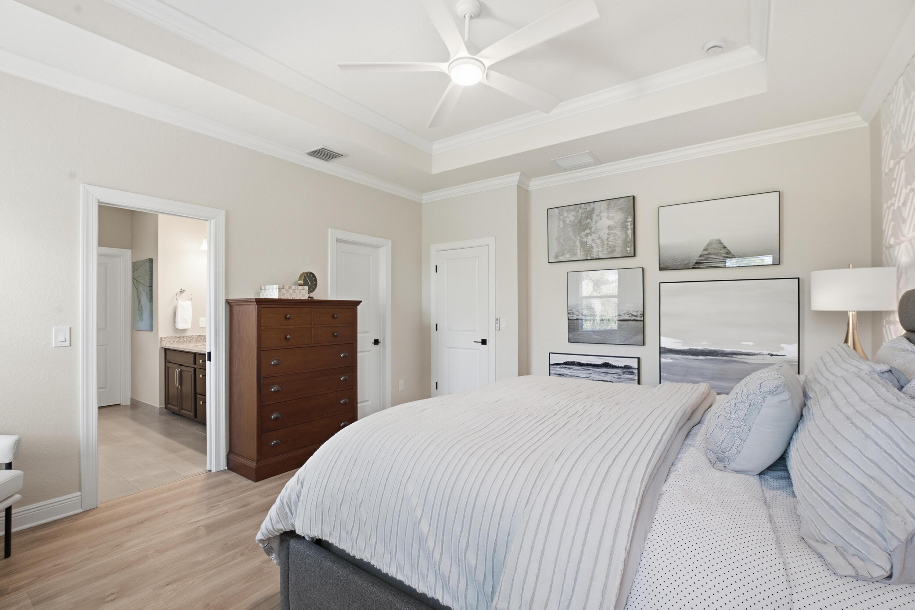 Primary bedroom with white striped bedding, a tray ceiling, coastal artwork, and an adjoining bathroom doorway.