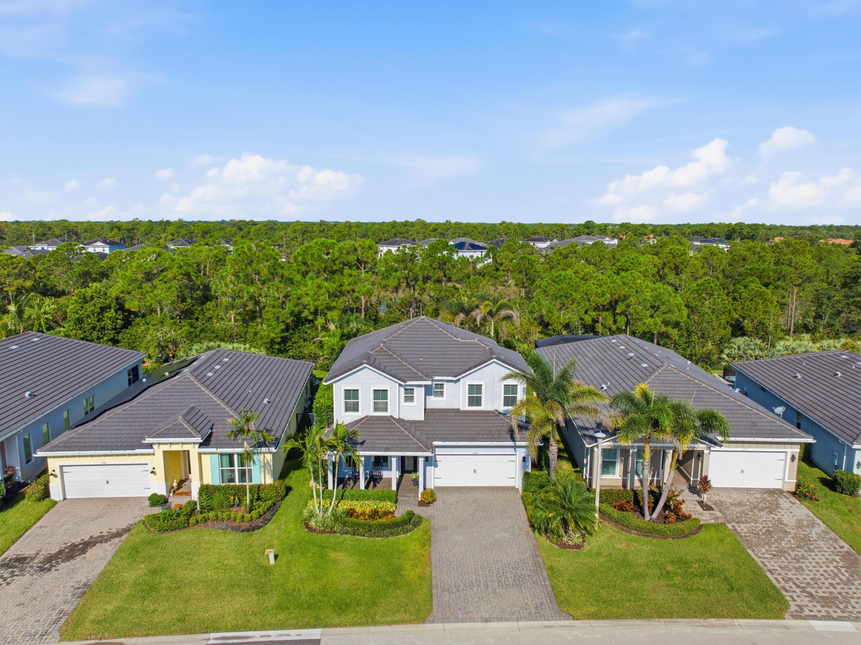 Aerial view of a light-colored home with a gray roof, front lawn, and driveway, surrounded by neighboring houses and trees.