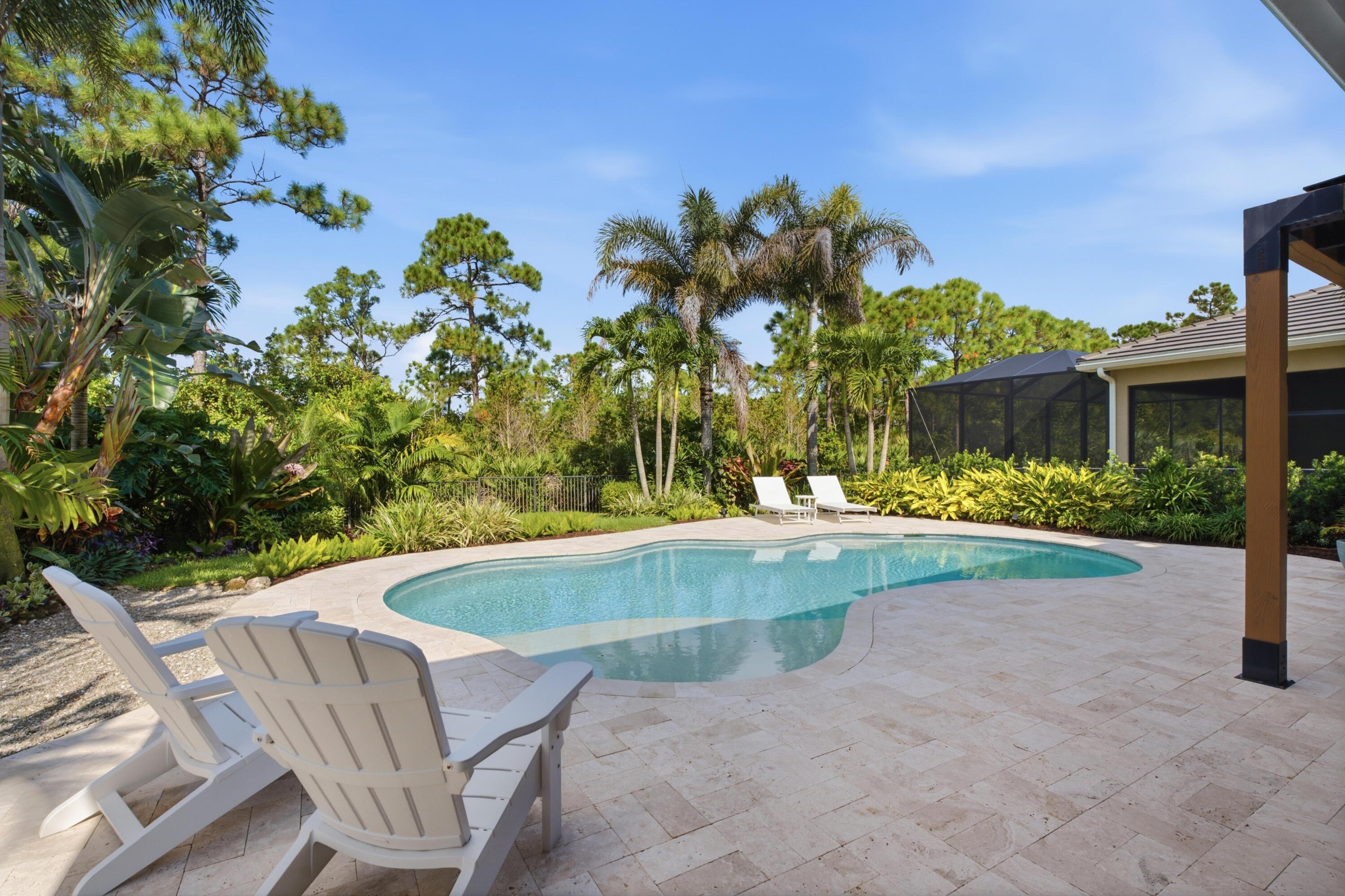 Backyard pool with a pale stone patio, white lounge chairs, and lush tropical landscaping.