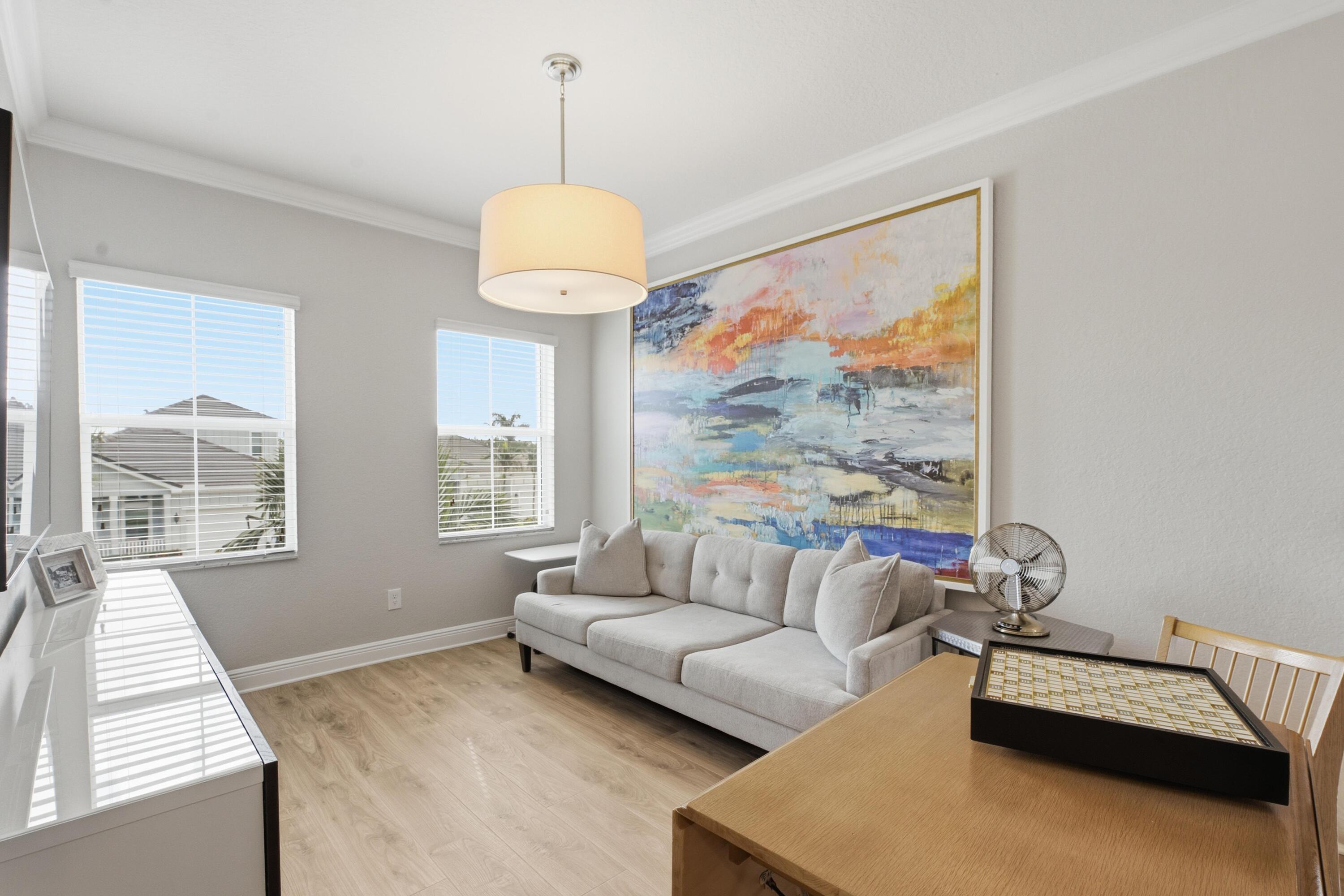 Furnished living room with a light gray sofa, colorful abstract painting, and large windows with white blinds.
