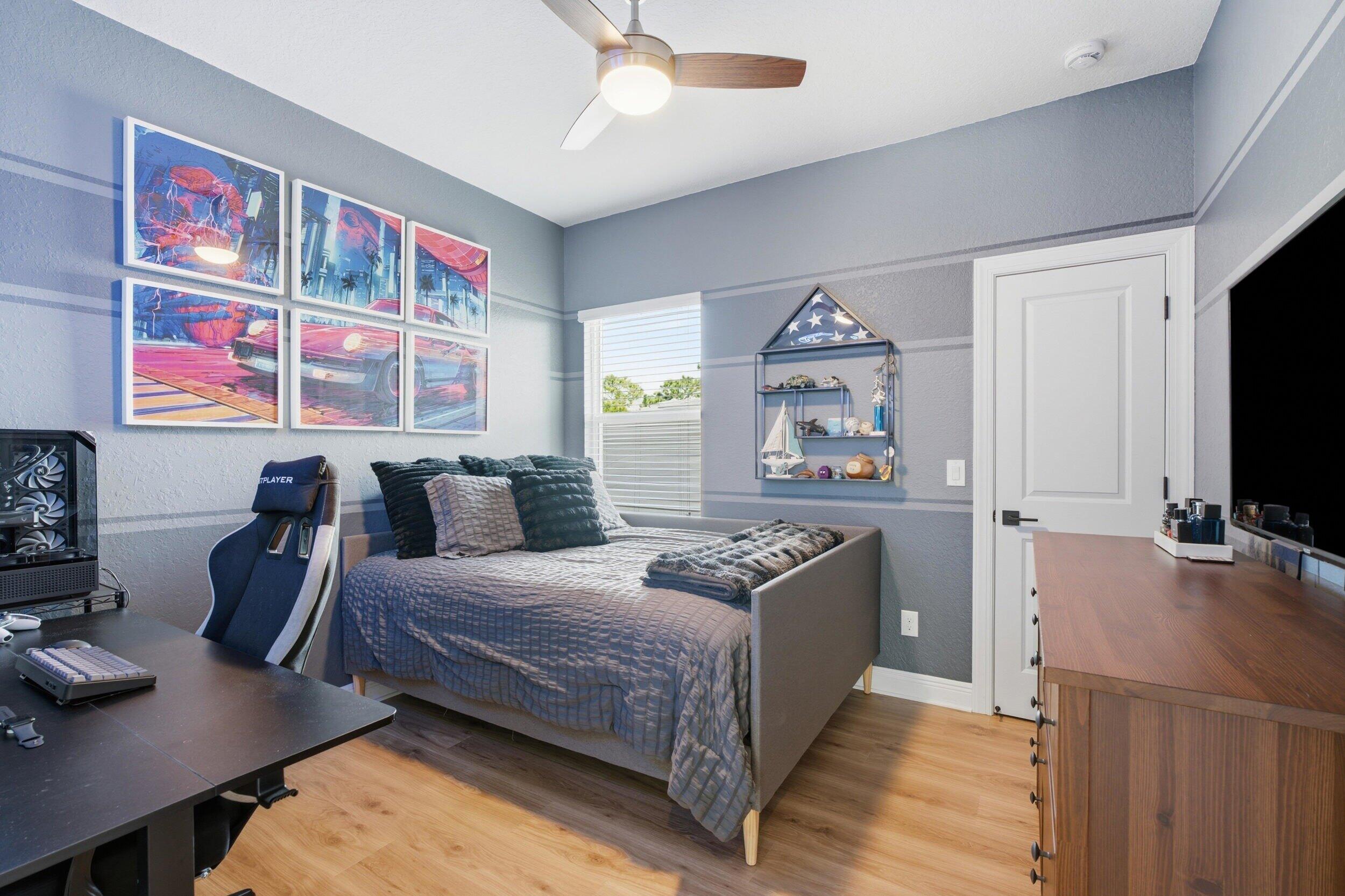 Bedroom with a gray bed, gaming setup, colorful framed artwork, and a wooden dresser.