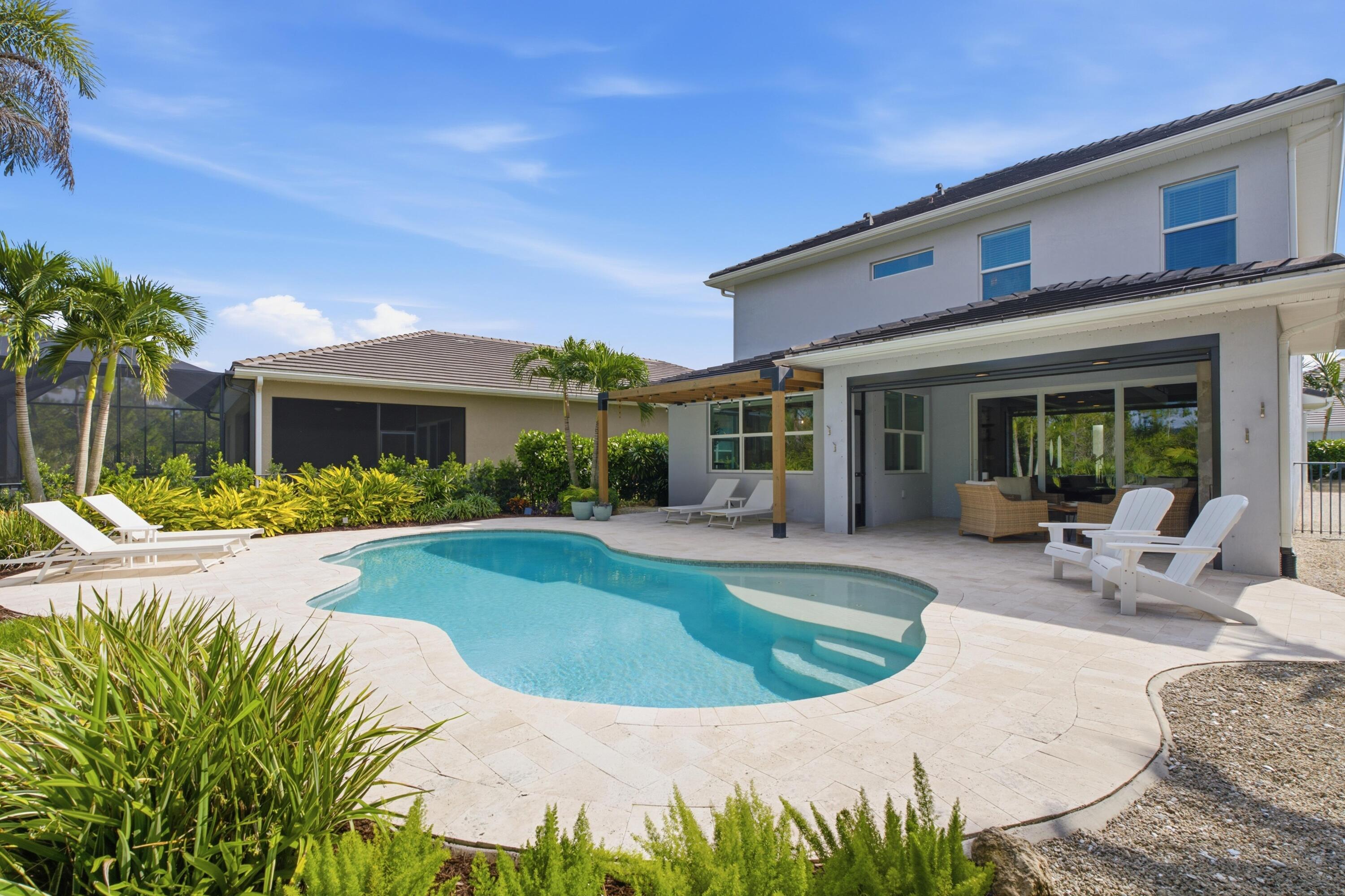 Backyard pool beside a two-story home, with a pale stone patio and tropical landscaping.
