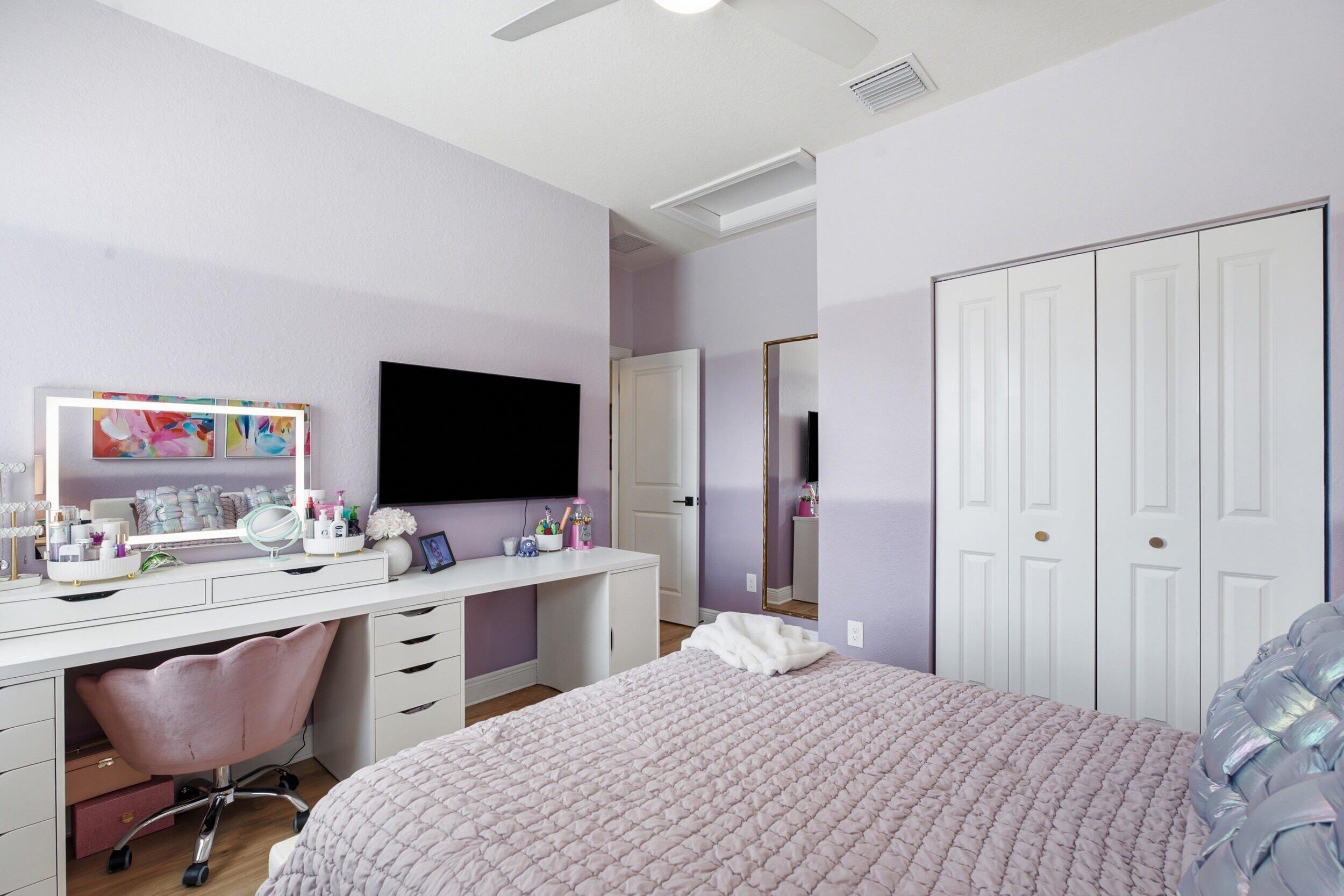 Bedroom with lavender walls, a pale quilted bed, white vanity, wall-mounted TV, and paneled closet doors.