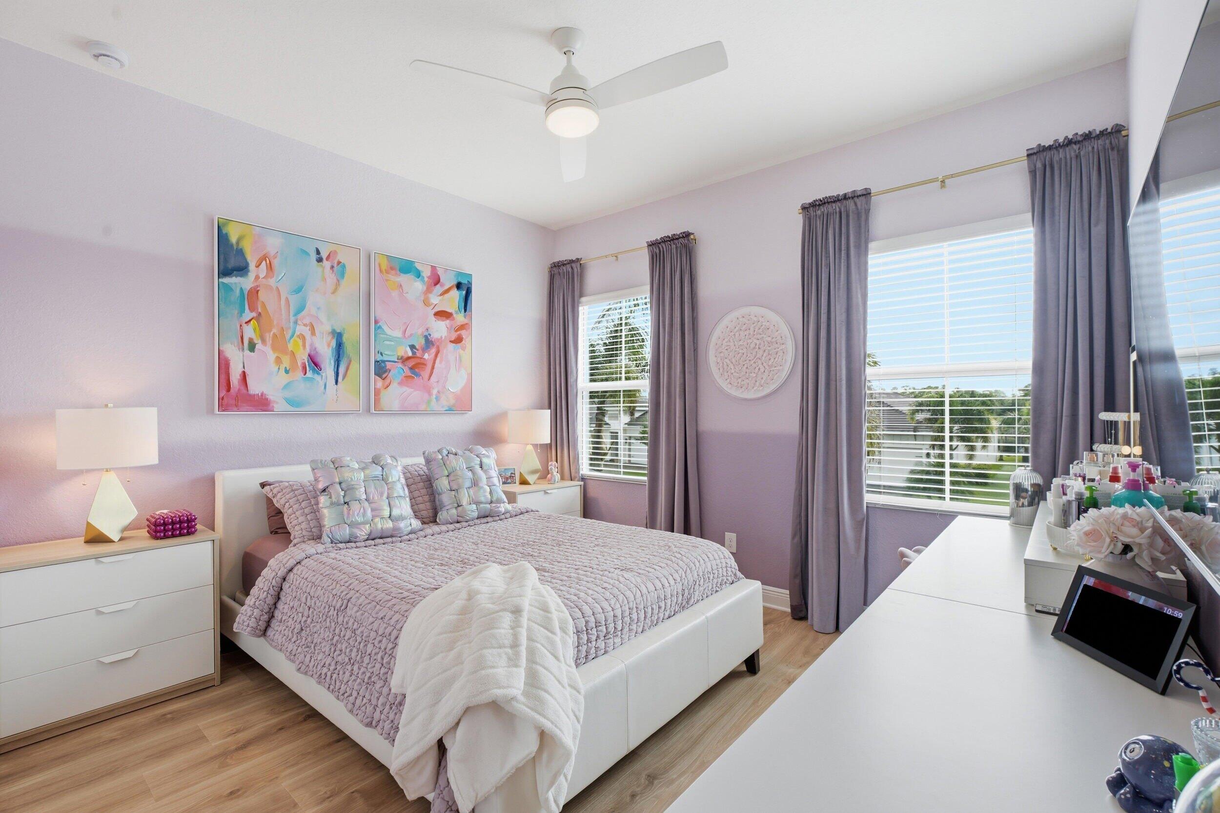 Furnished bedroom with lavender bedding, colorful abstract artwork, and large curtained windows.