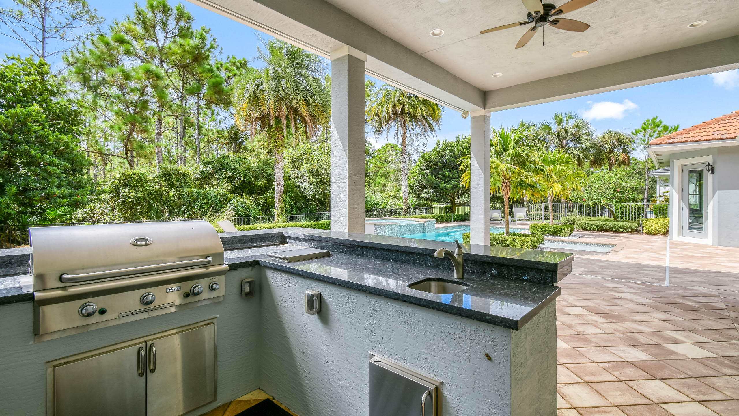 Covered outdoor kitchen with built-in grill and sink overlooking a pool and tropical landscaping.