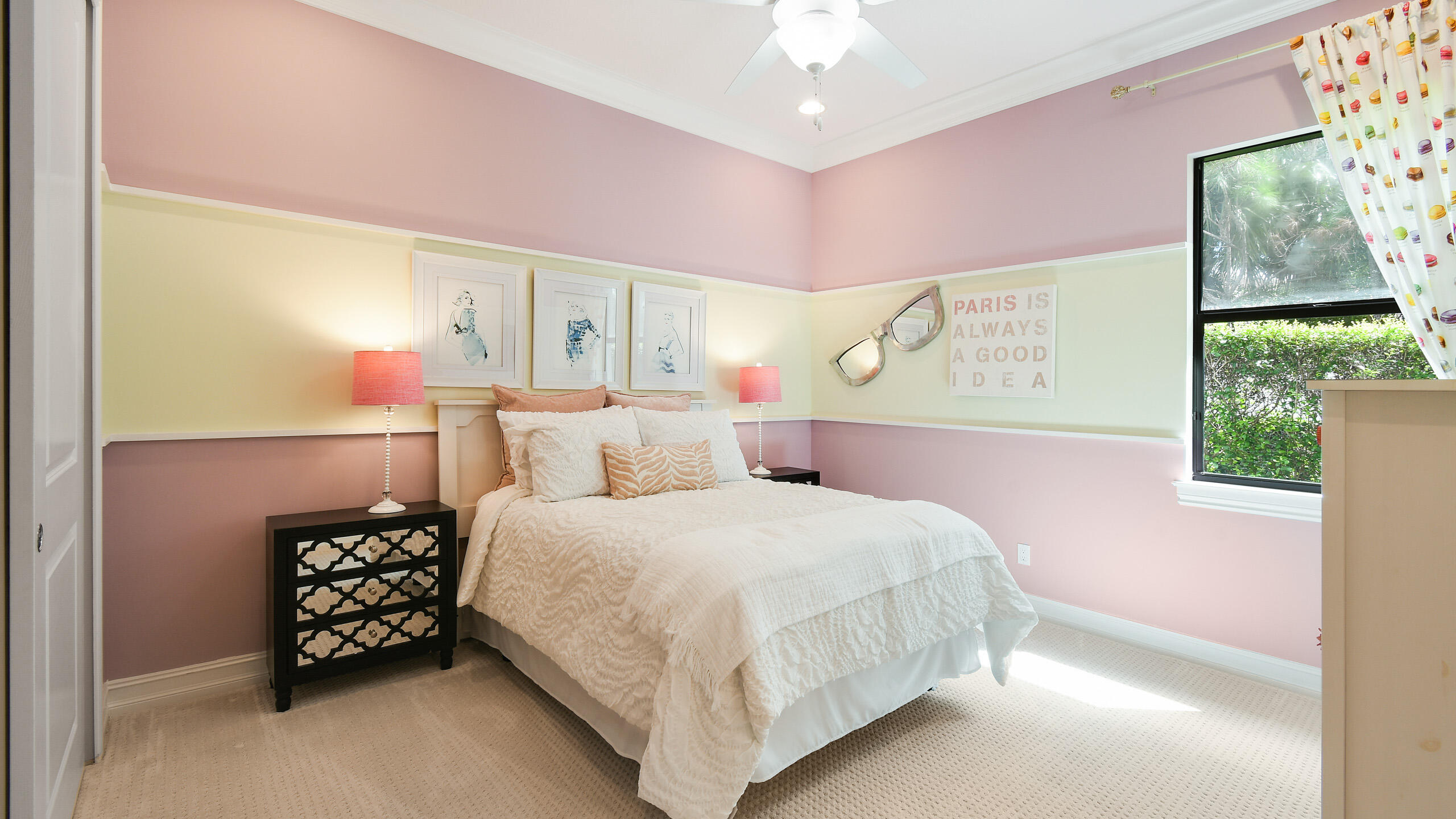 Furnished bedroom with a white bed, pink and yellow walls, and a large window overlooking greenery.