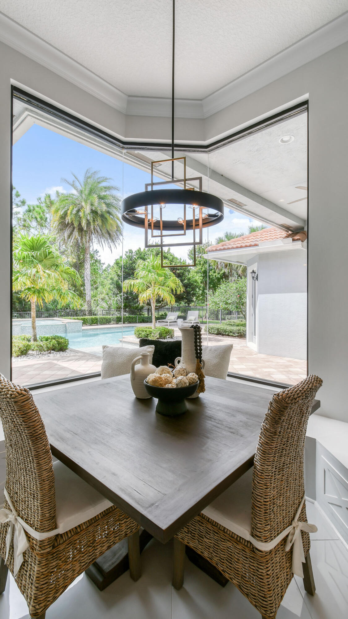 Dining area with woven chairs and corner windows overlooking a pool and tropical landscaping.