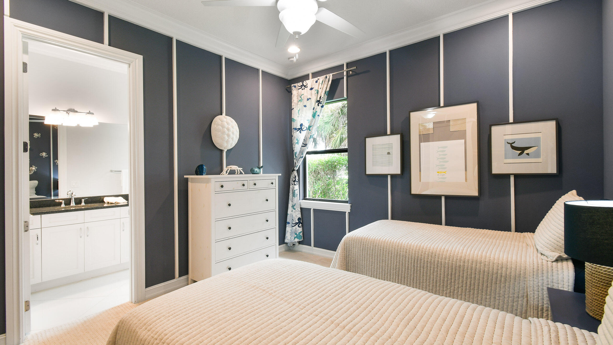 Bedroom with two cream quilted beds, dark paneled walls, framed fish art, and an adjoining bathroom vanity.