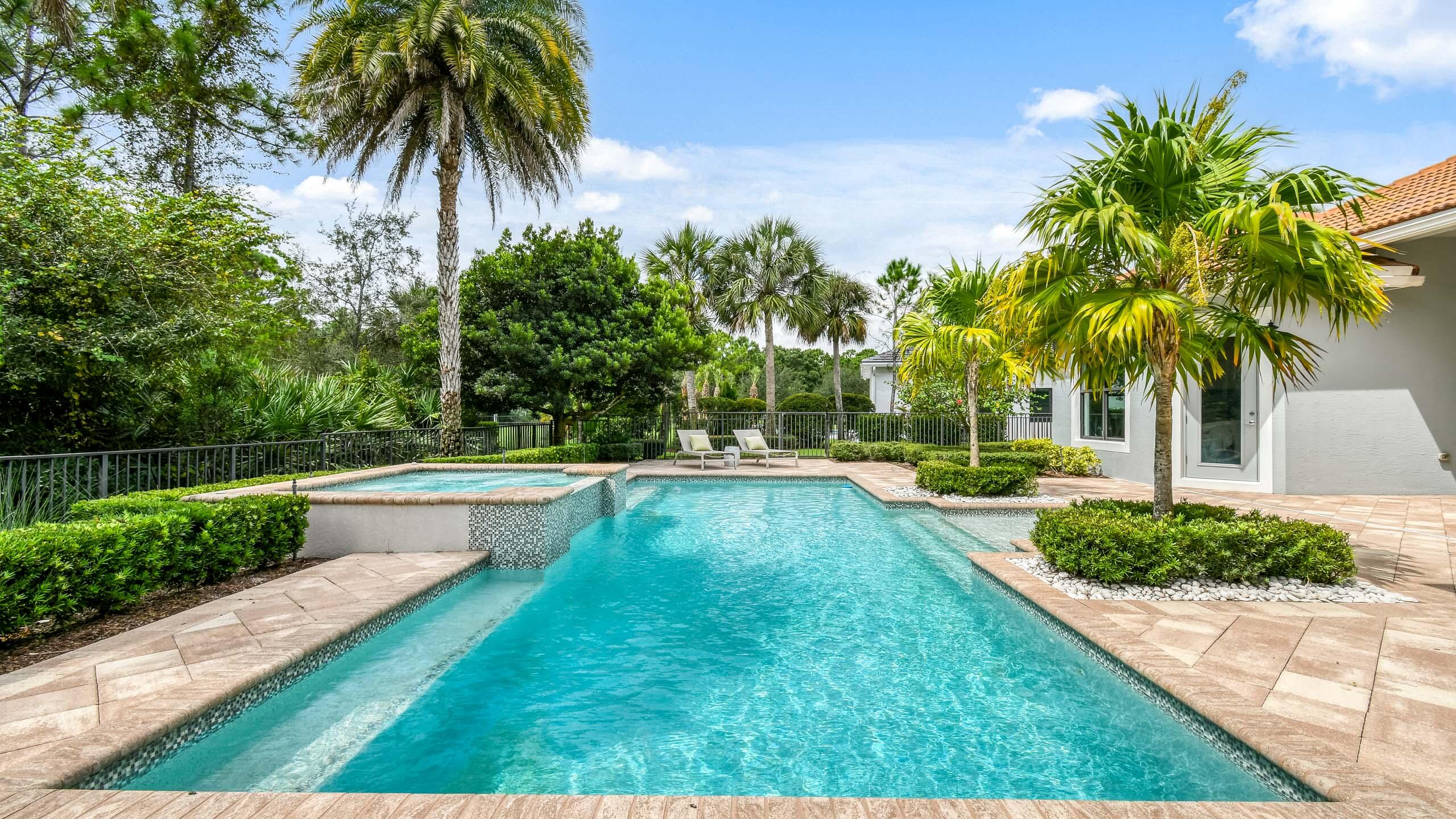 Backyard swimming pool with raised spa, tropical palms, and paved patio beside a white stucco home.