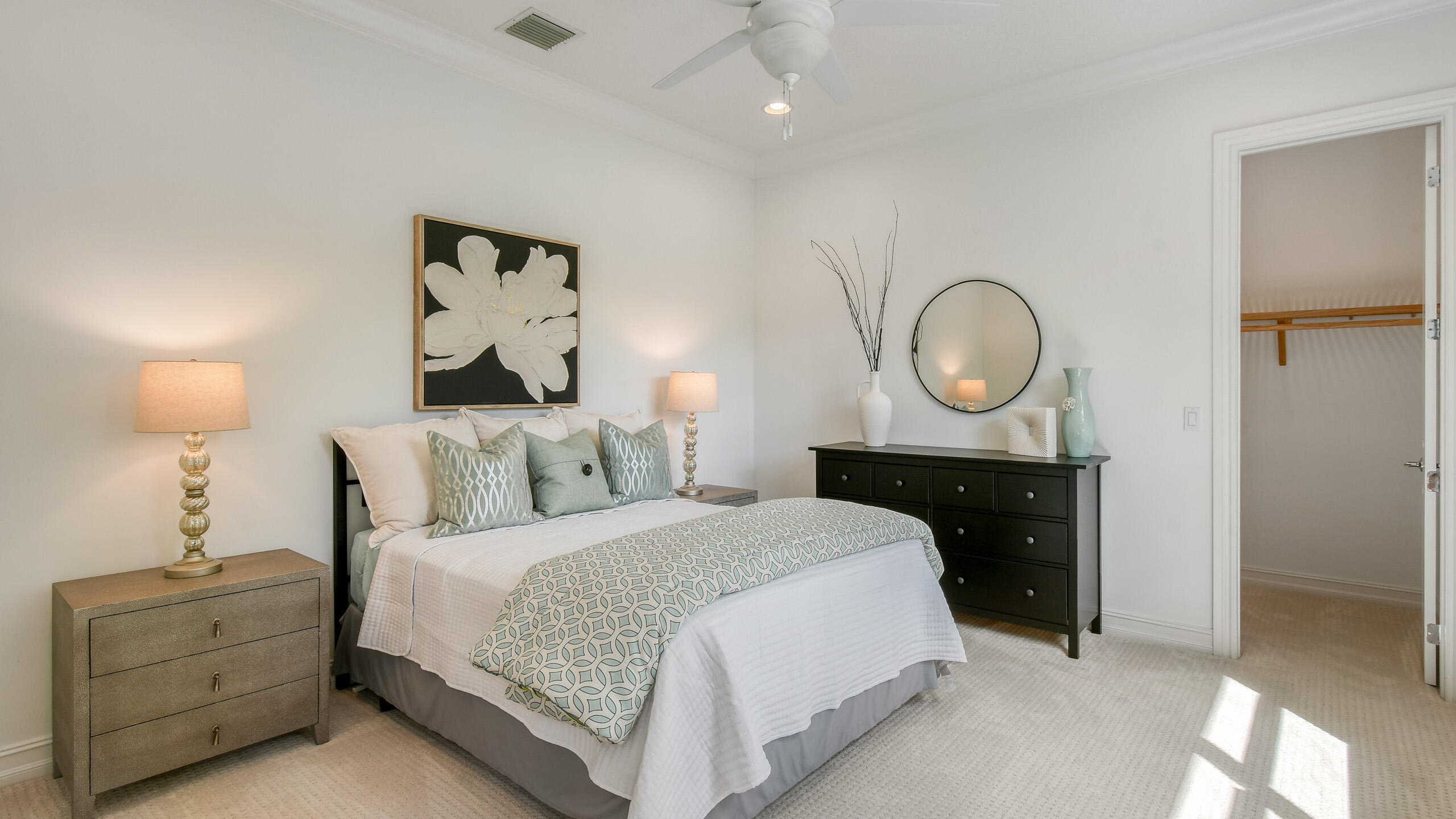 Staged bedroom with pale blue patterned bedding, a black dresser, and sunlight across the carpet.