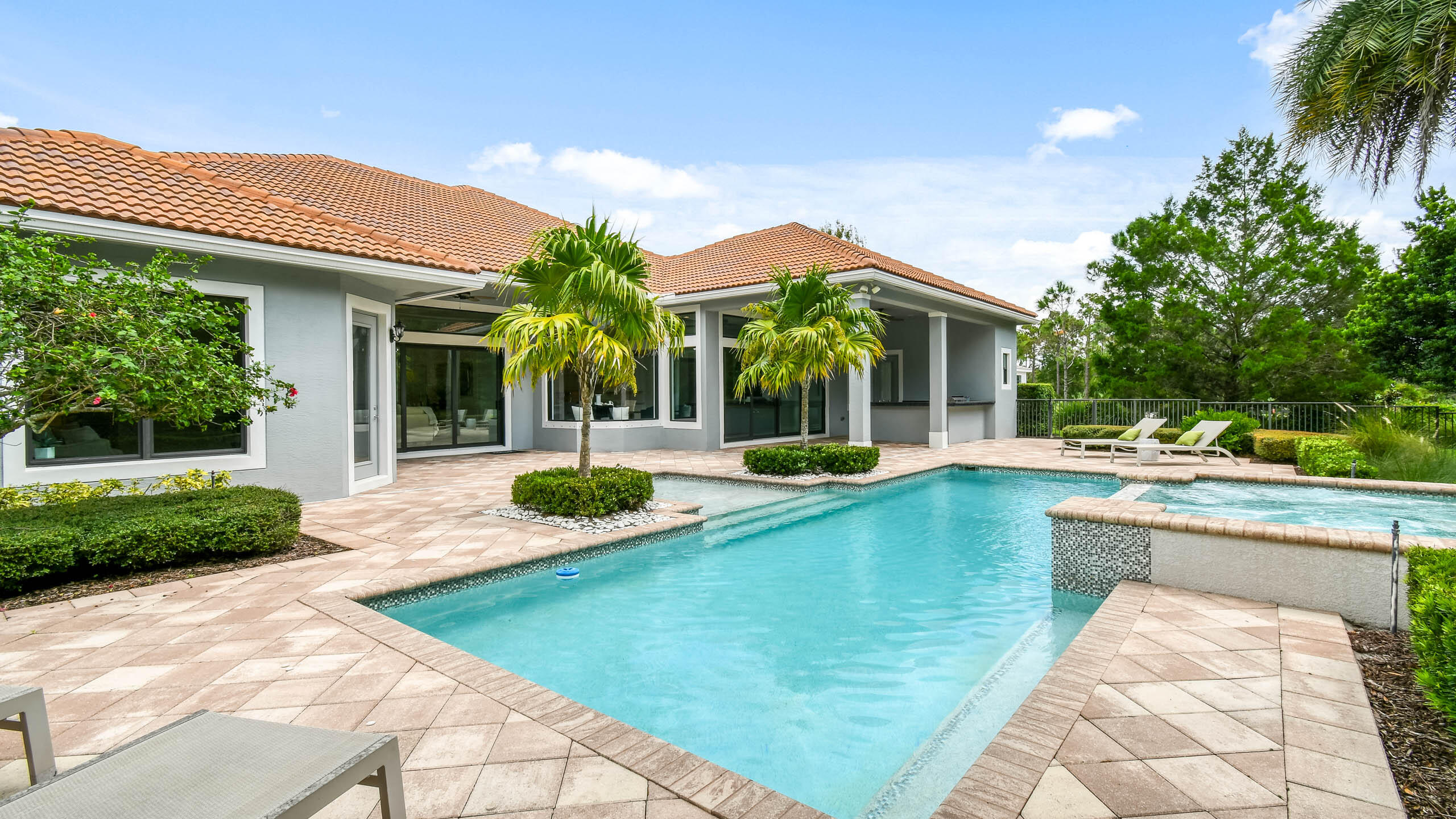 Backyard swimming pool and raised spa beside a gray home, with palm trees and a paved patio.