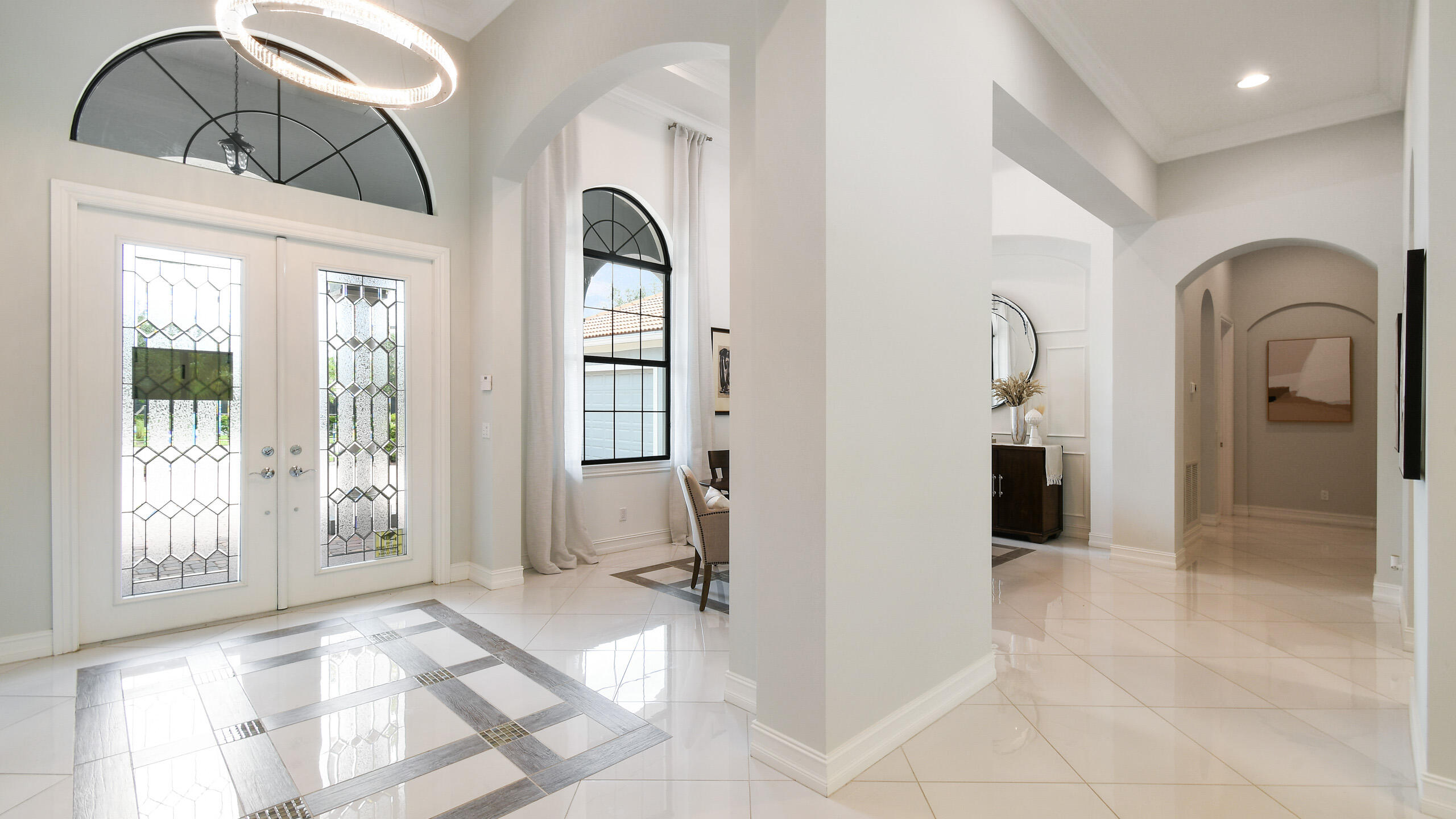 Bright foyer with decorative glass entry doors, arched windows, and glossy tile flooring with gray inlay.